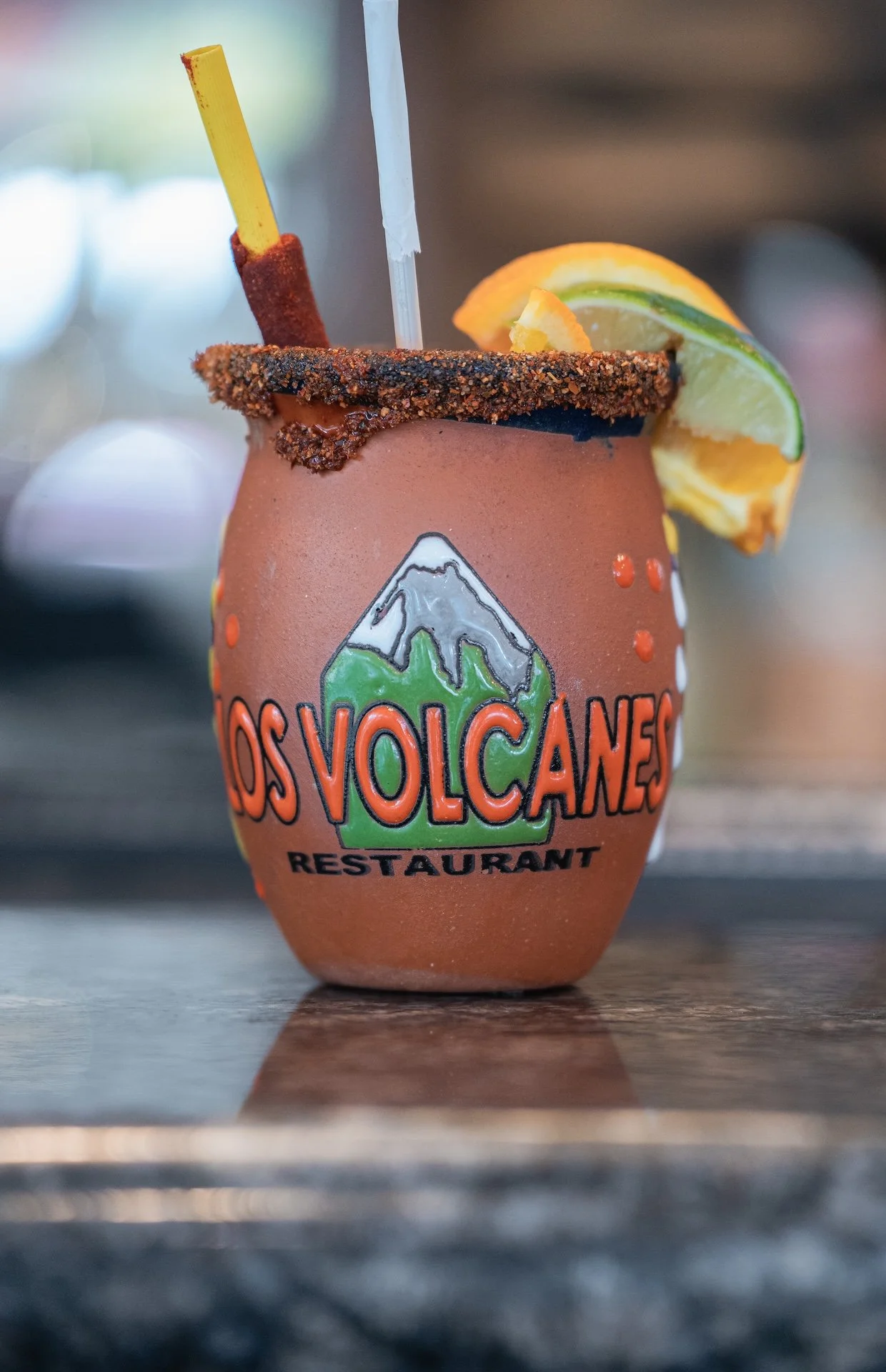 A cocktail in a cup with the logo of a volcano and the words 'Los Volcanes Restaurant'. The drink is garnished with slices of lemon and lime, and has a chili-rimmed glass with a drink stirrer.