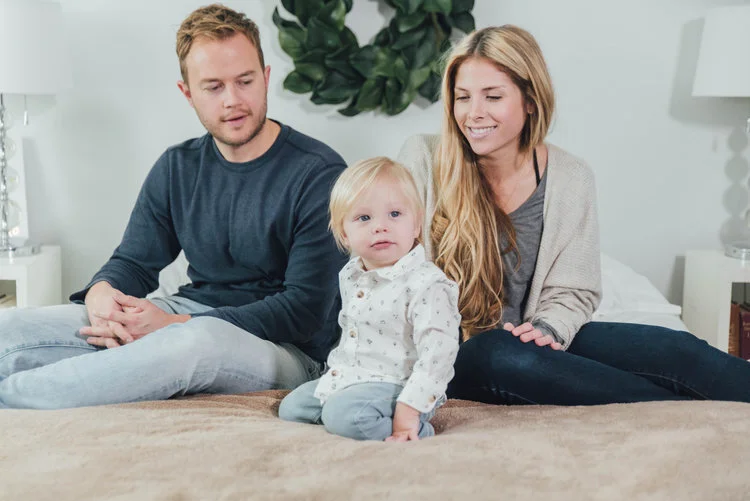 A family sitting on a bed with a toddler in the center and parents on either side.