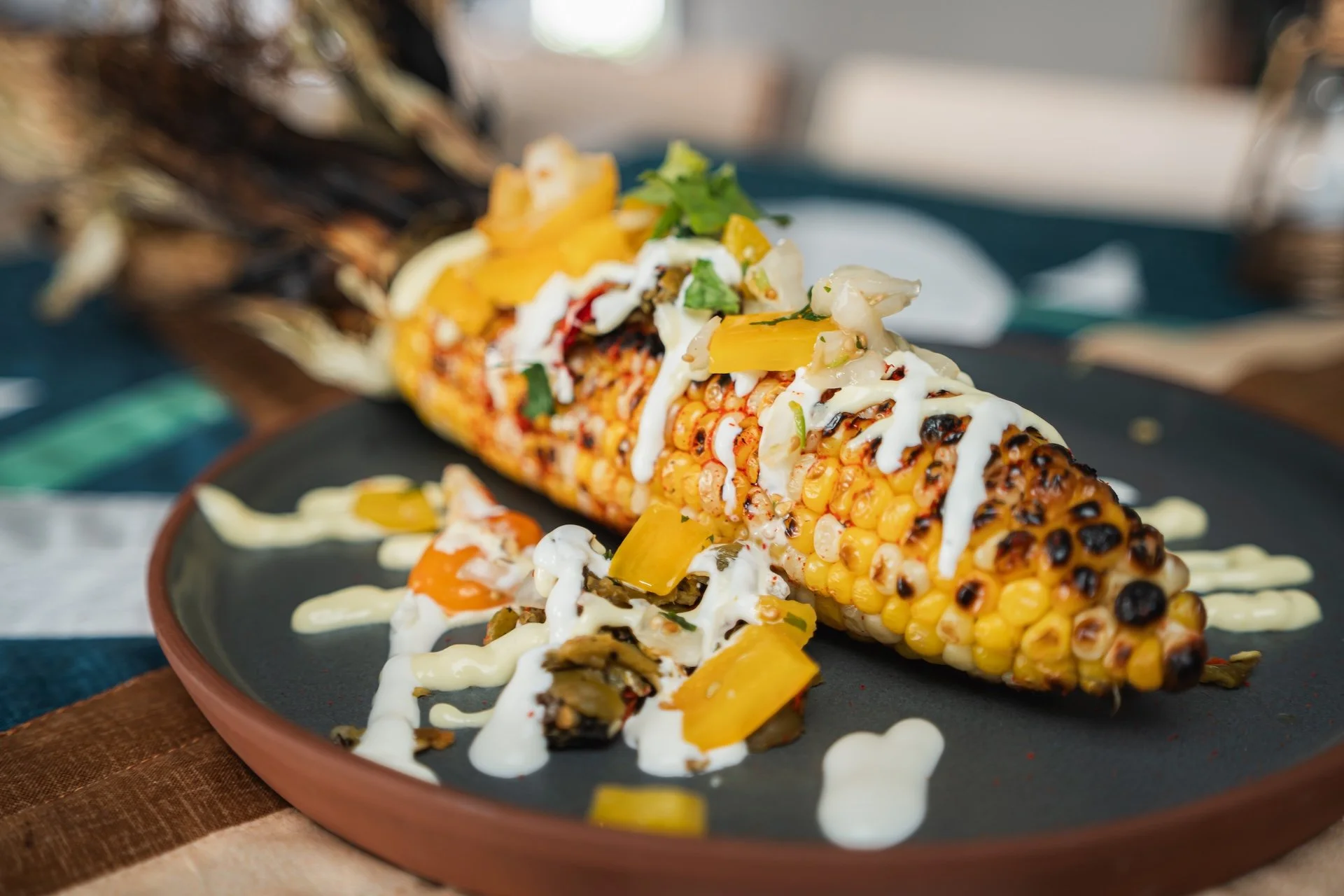 Grilled corn on the cob topped with white sauce, chopped green herbs, and yellow peppers, served on a black plate with additional sauce and toppings, on a wooden table.