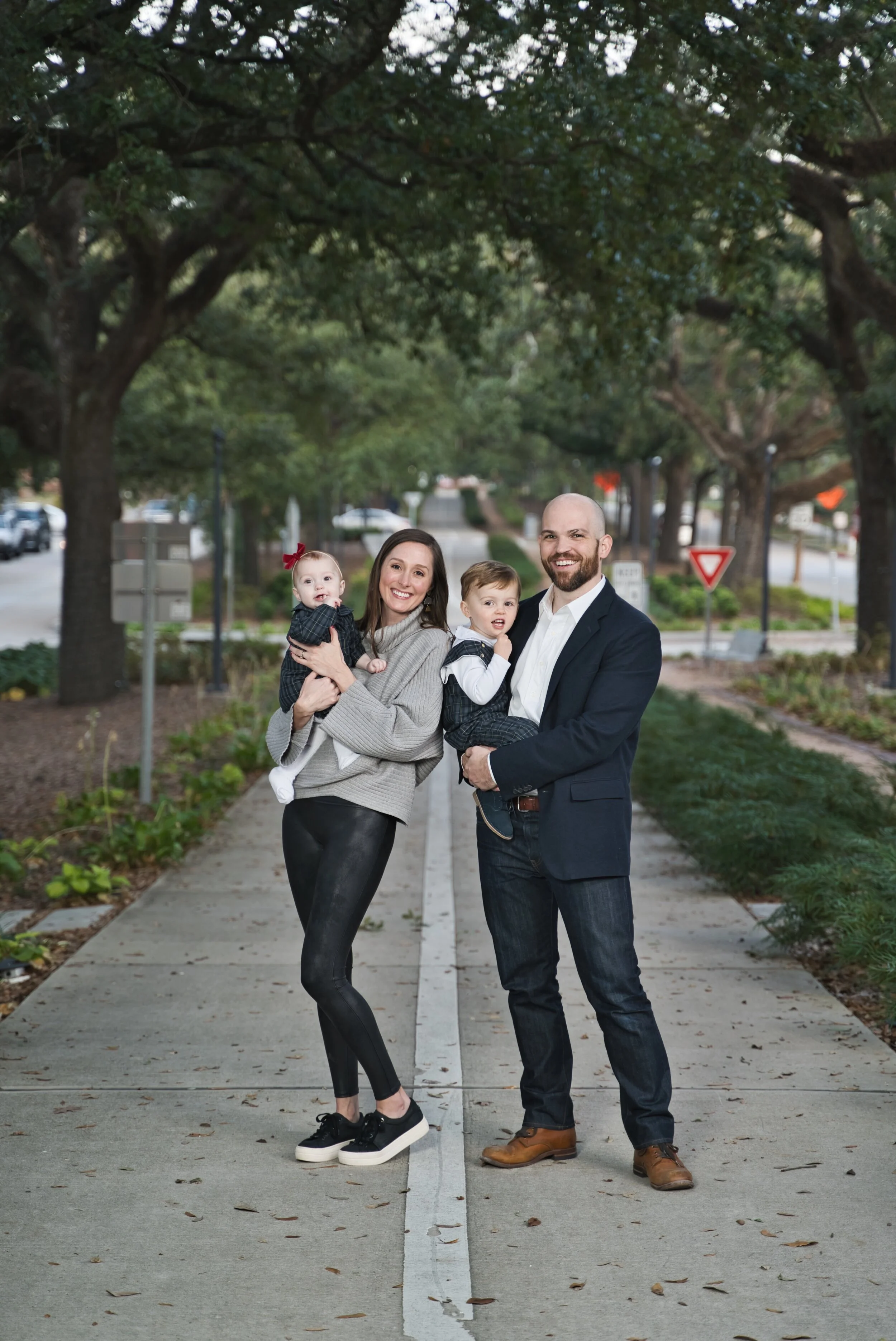 Family of four posing outdoors, with two adults holding two young children, standing on a sidewalk lined with trees.