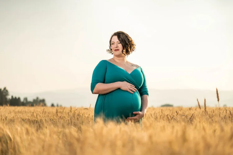 Pregnant woman in teal dress standing in a wheat field, holding her belly, with a serene landscape in the background.