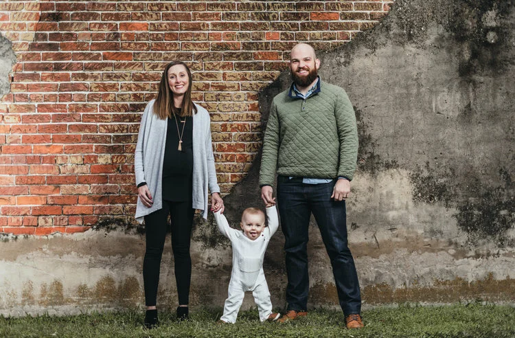 A family of three posing in front of a weathered brick wall.