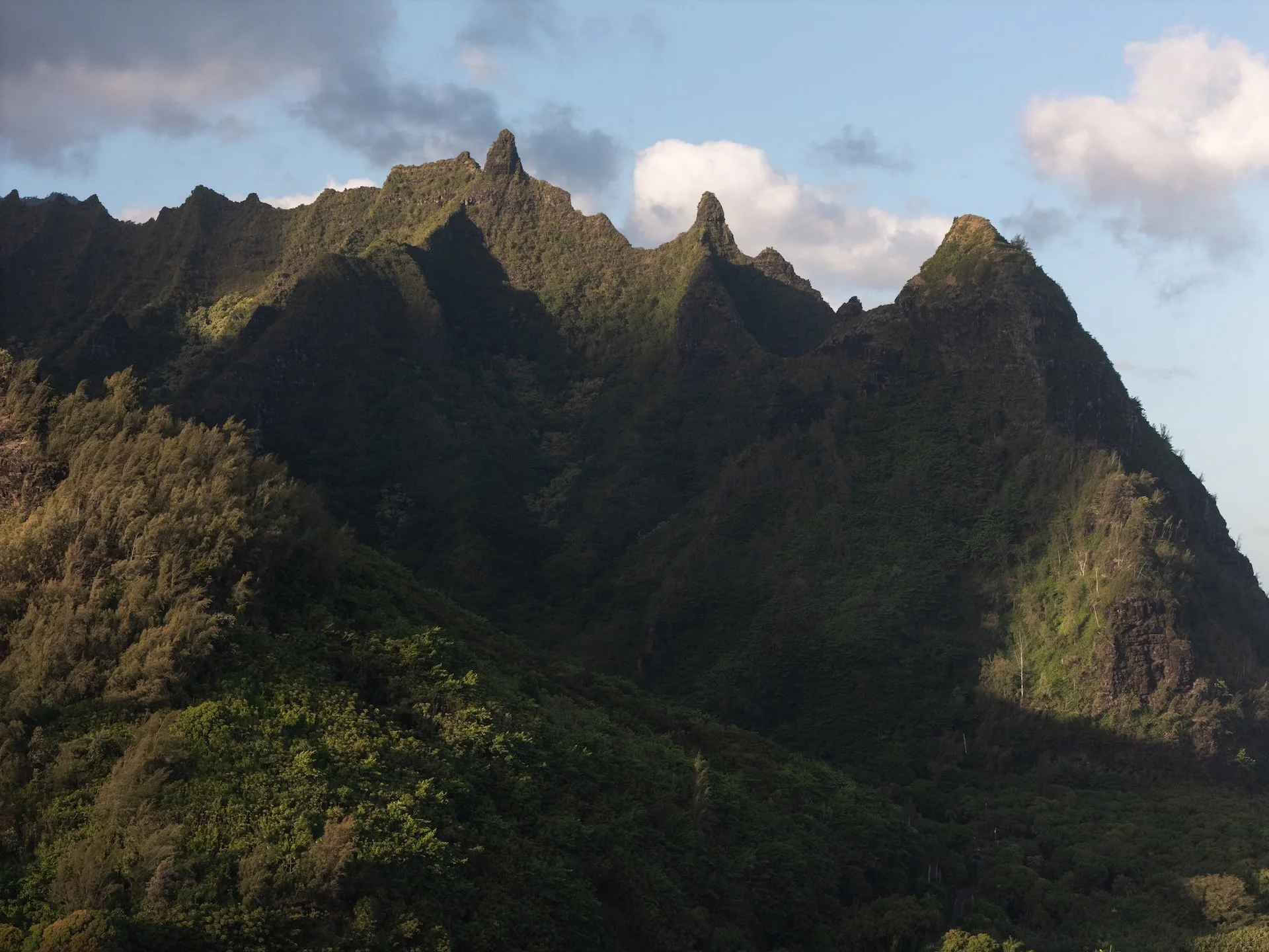 Sunlight illuminates green, jagged mountain ridges under a partly cloudy sky.