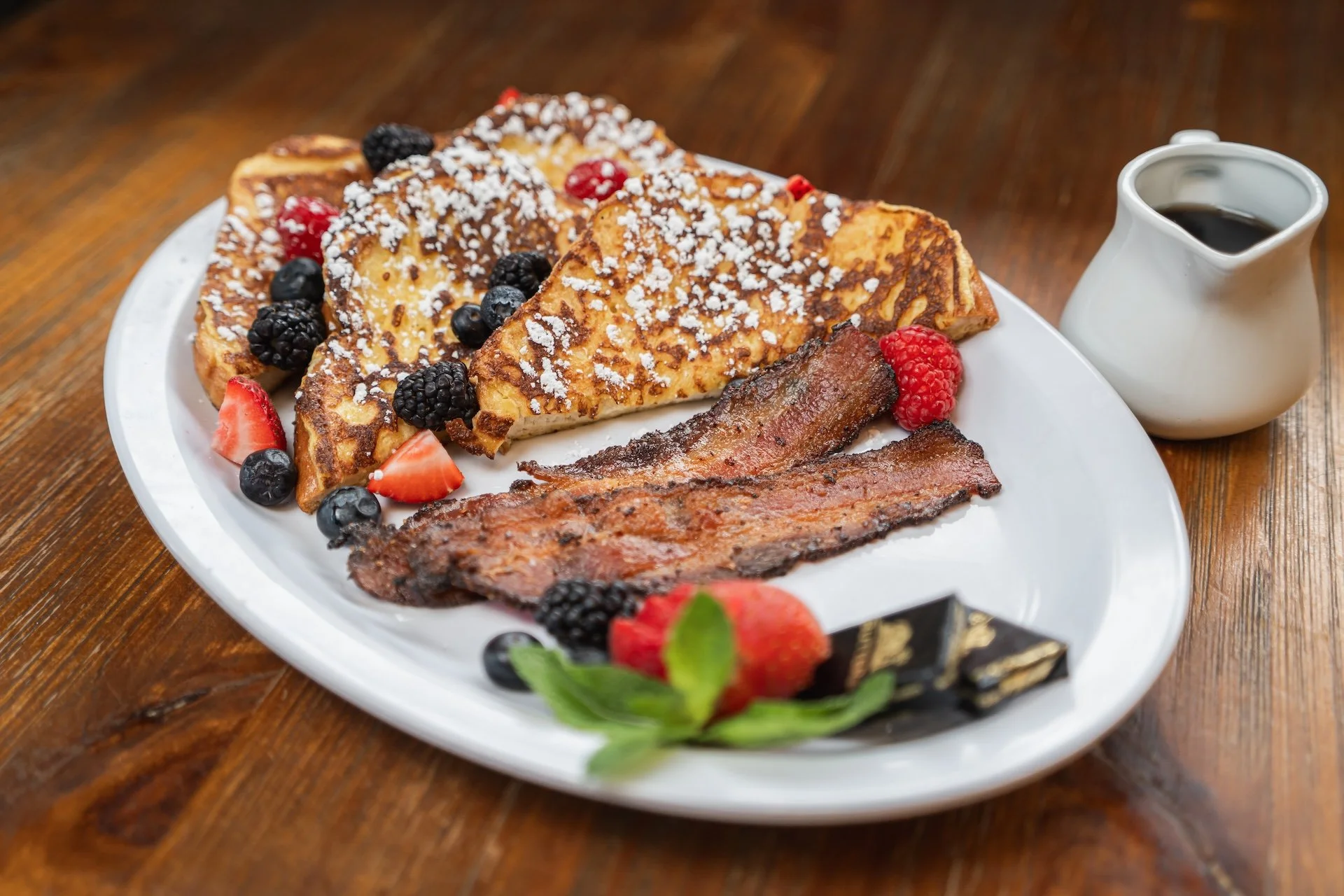 Plate with French toast topped with powdered sugar, fresh berries, and slices of bacon, alongside a small pitcher of syrup, all on a wooden table.