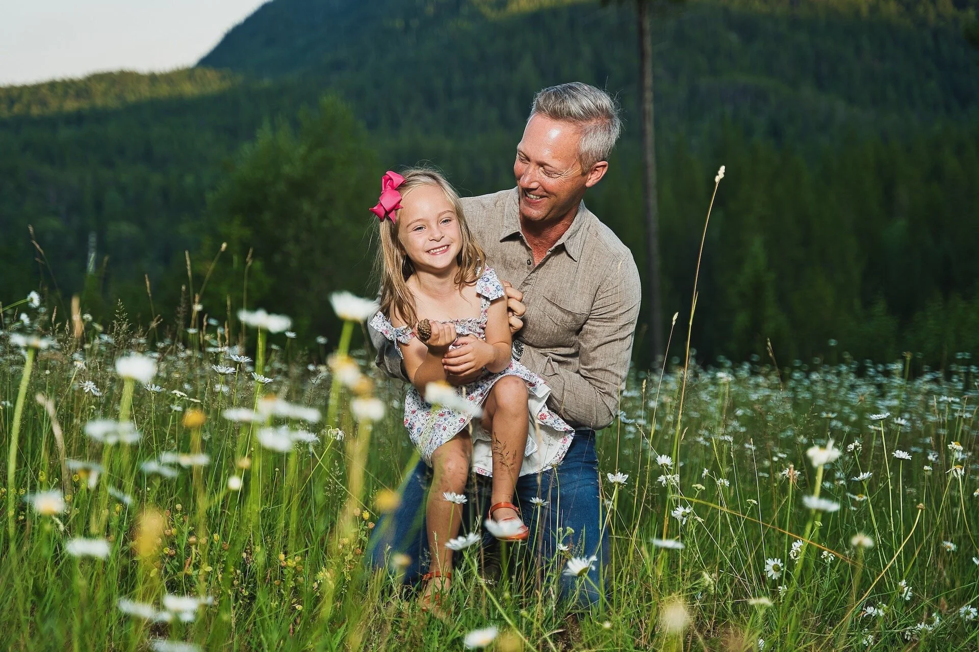 Man and young girl smiling in a field of flowers with mountains in the background.
