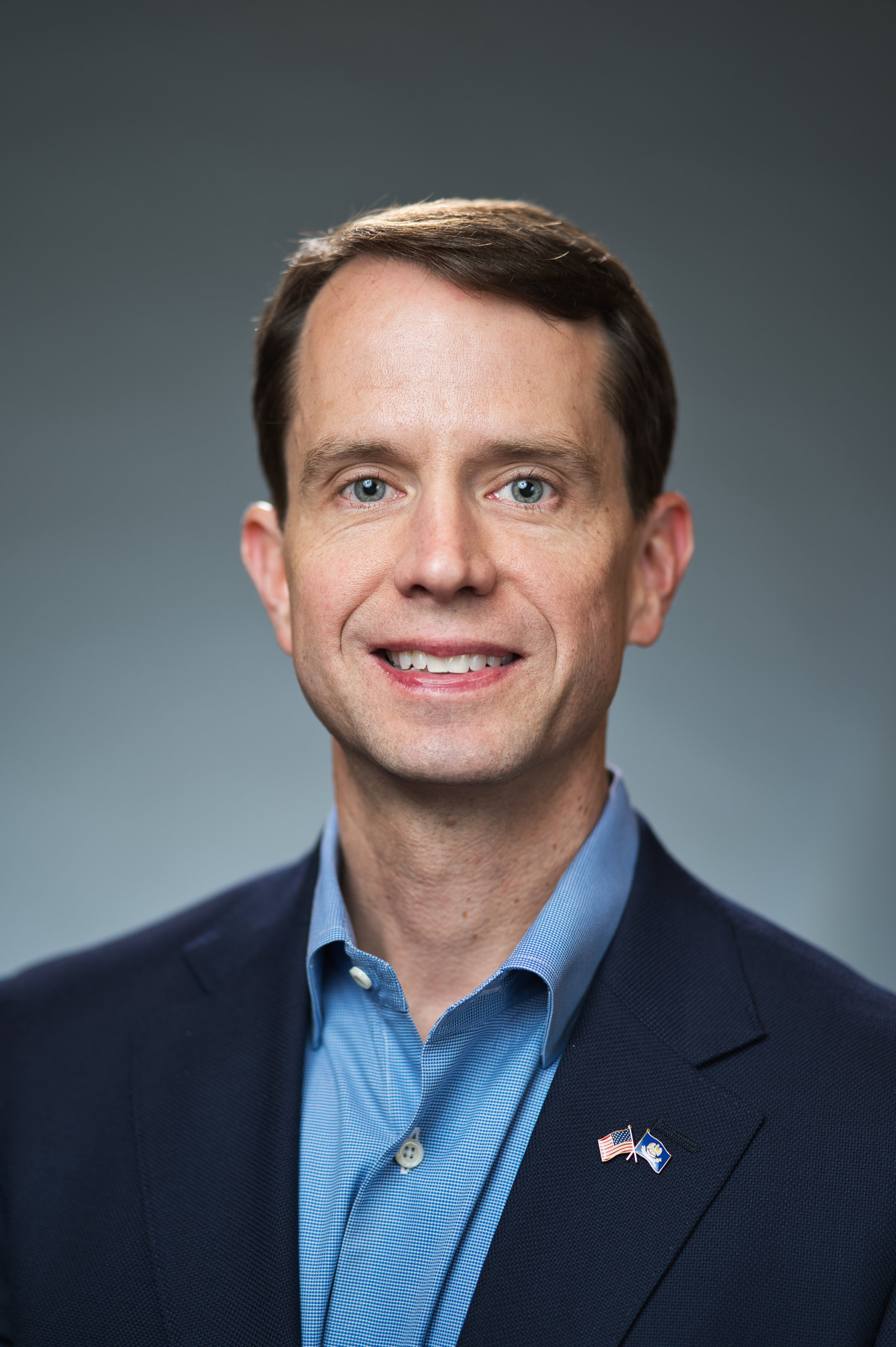 Man in a blue shirt and navy blazer with a small American flag pin.