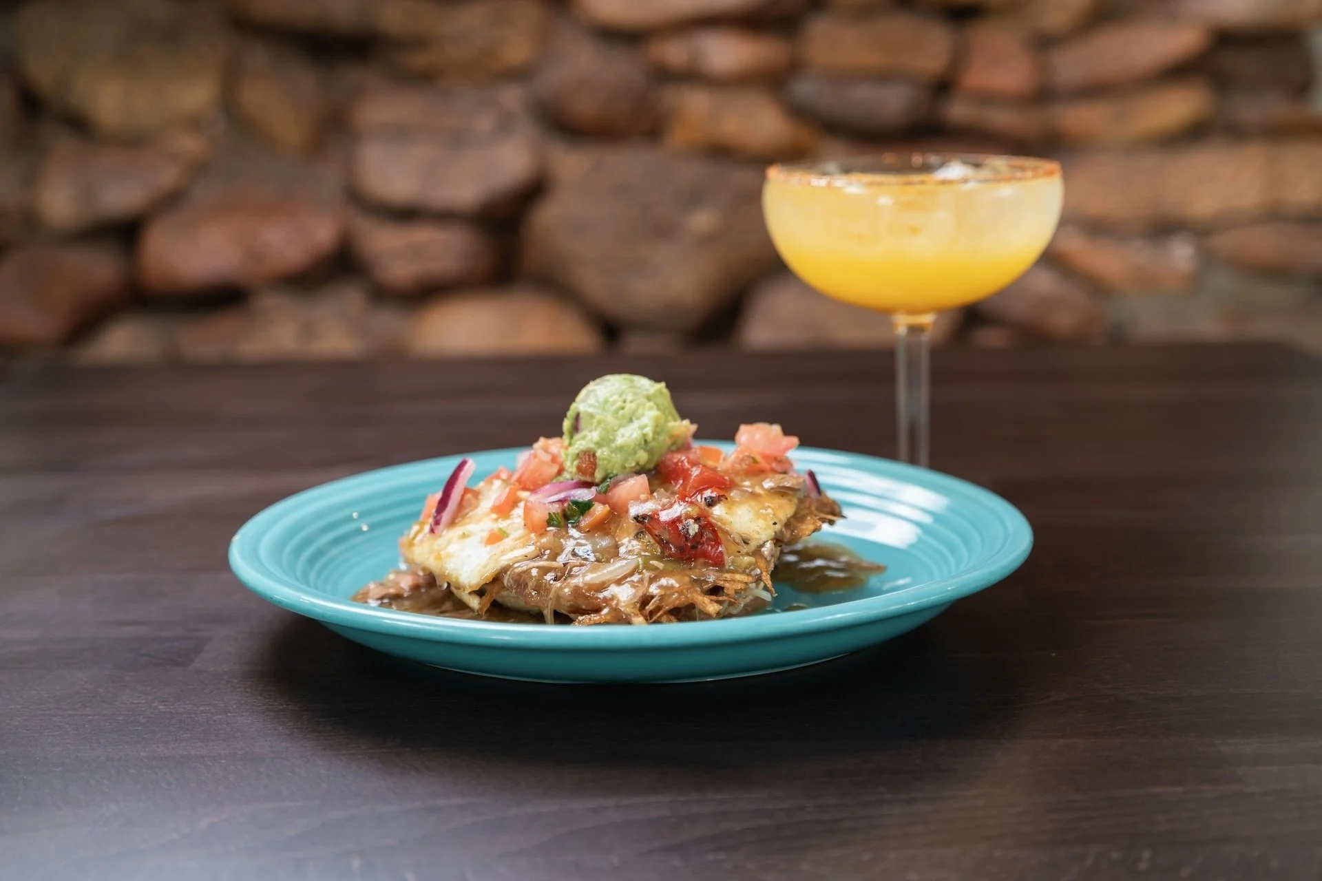 A plate of Mexican dish topped with guacamole and pico de gallo, served with a cocktail in a coupe glass as background.