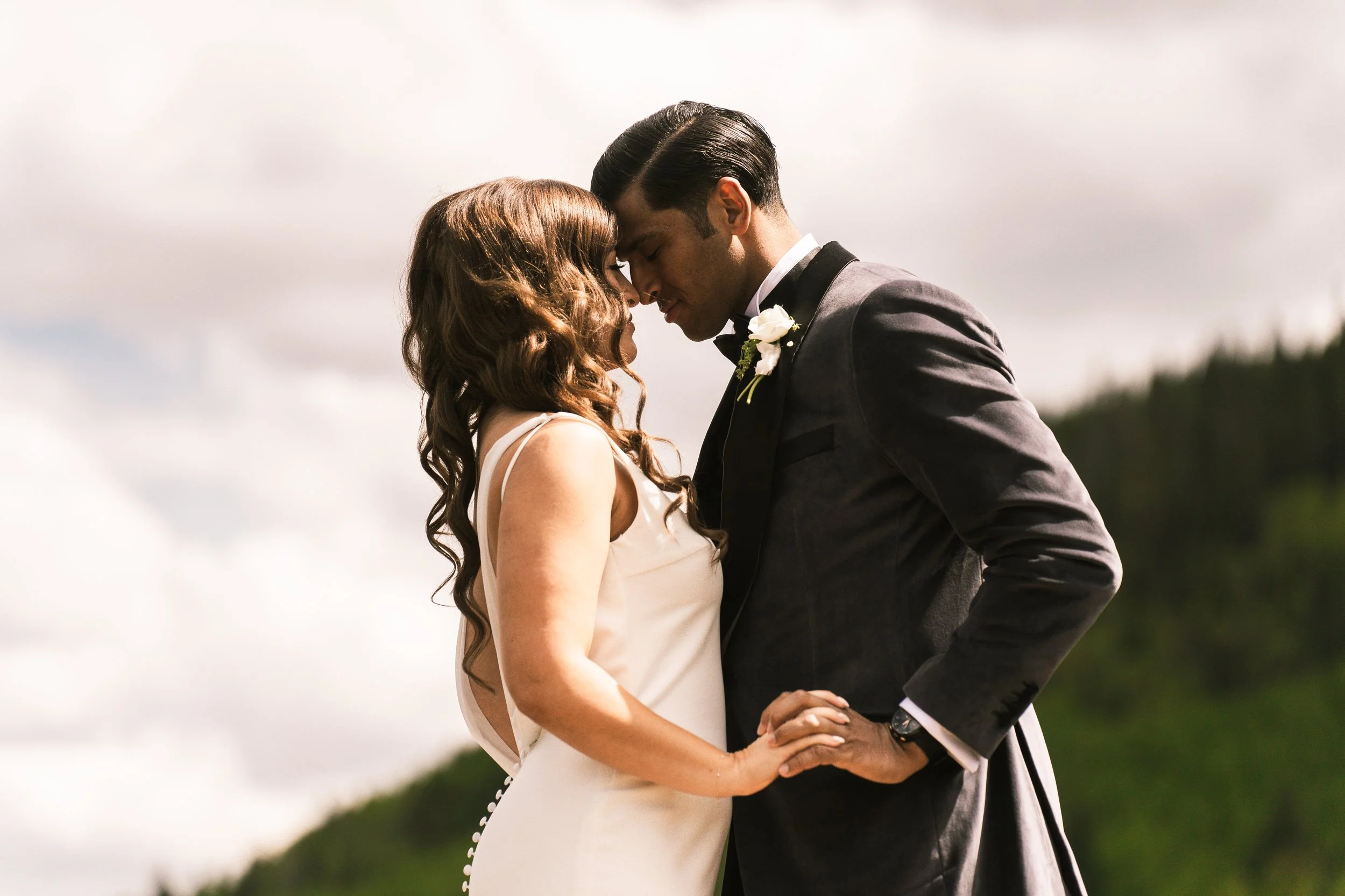 A couple in formal attire embracing outdoors with a natural landscape and cloudy sky in the background.
