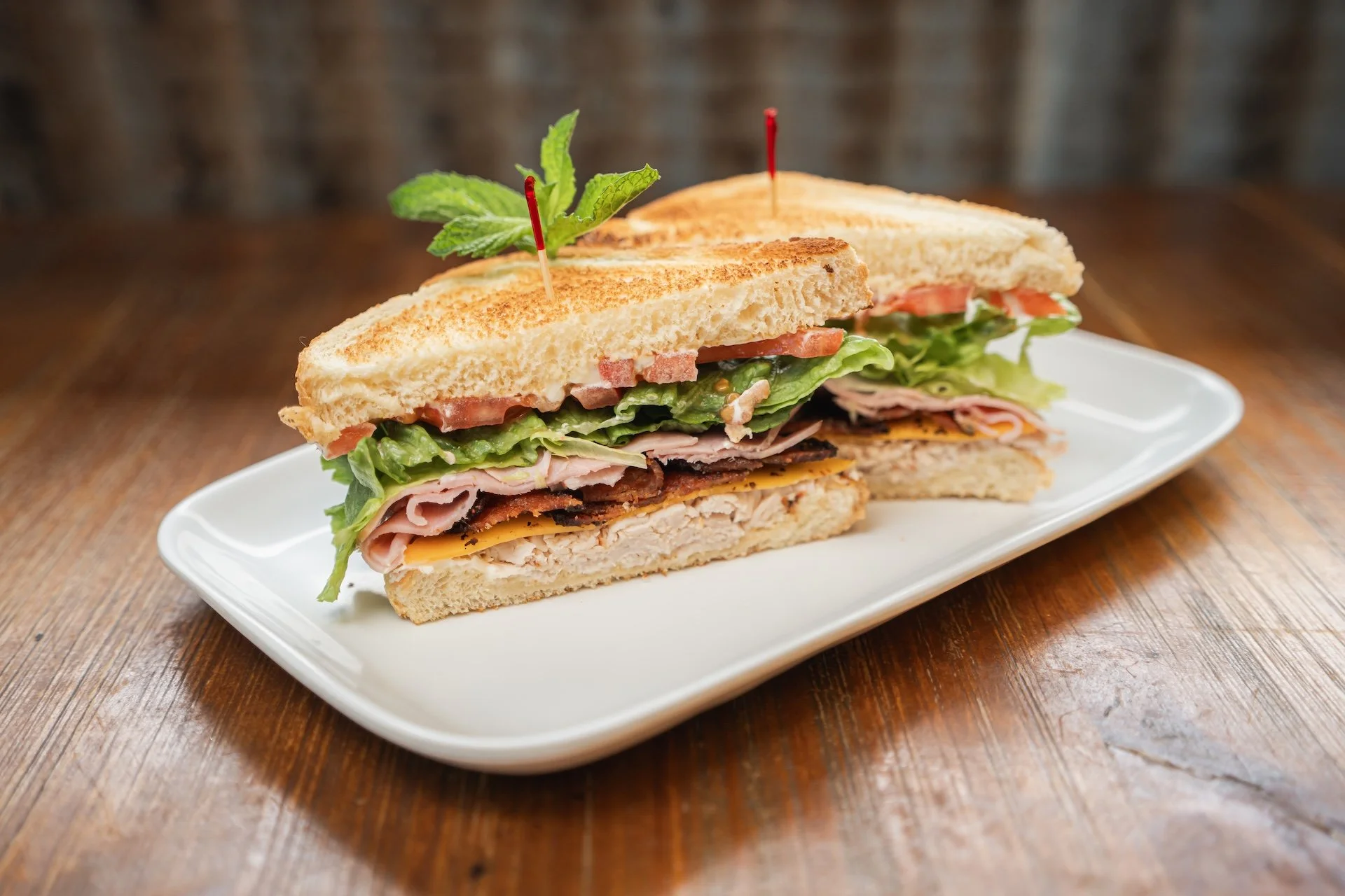 Two halves of a breakfast sandwich on a white plate, with toasted bread, lettuce, tomato, turkey, ham, cheese, and garnished with a sprig of mint and toothpicks.