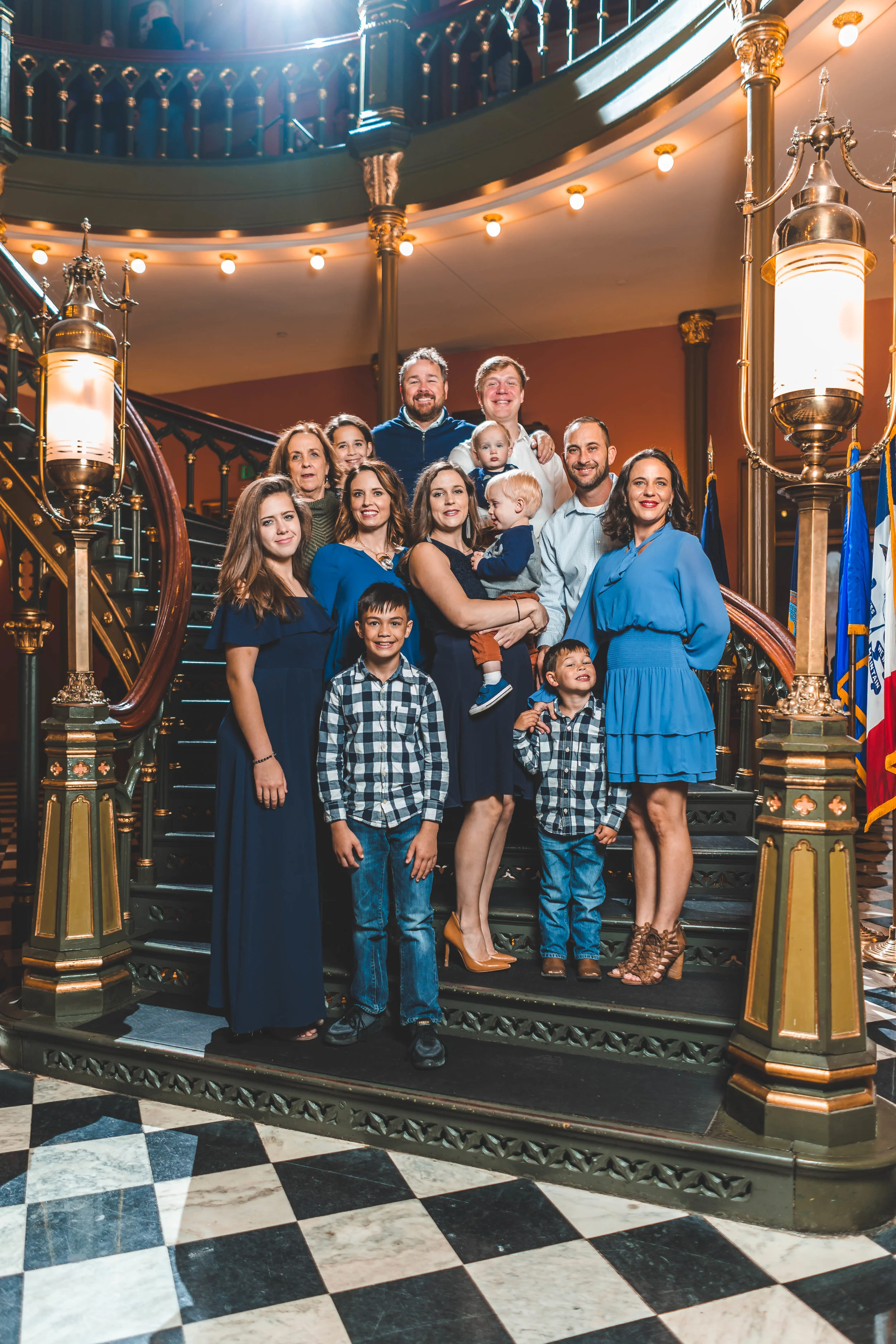 A group of people, including adults and children, posing together on an ornate staircase with golden railings indoors. They are dressed in formal and semi-formal attire, with some wearing blue outfits. The setting includes detailed architecture and d