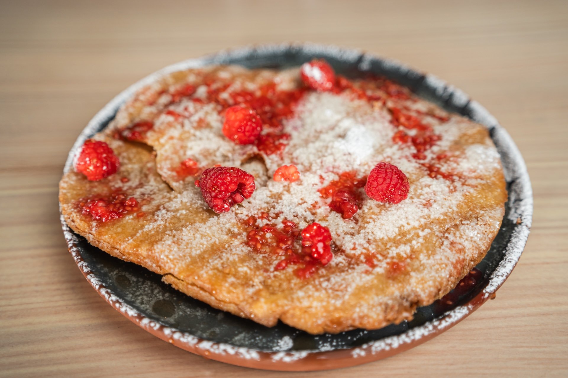 Fried bread topped with fresh raspberries, powdered sugar, and strawberry syrup, served on a black plate on a wooden table.