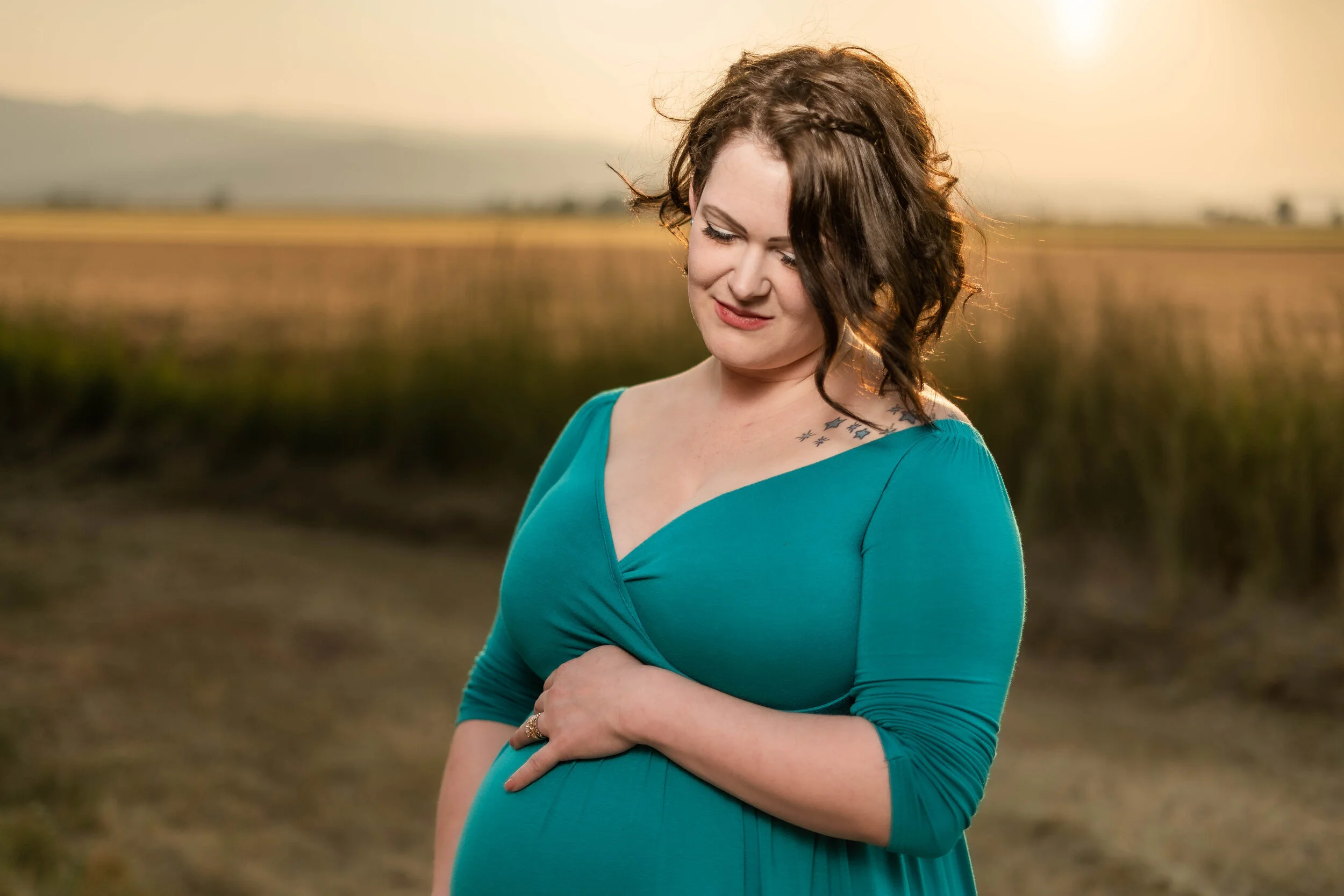 Pregnant woman in teal dress standing in a field at sunset, holding her belly and looking down with a serene expression.