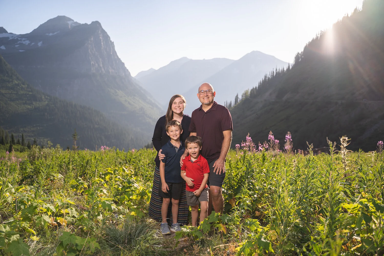 Family of four posing in a lush field with mountains in the background during daytime.