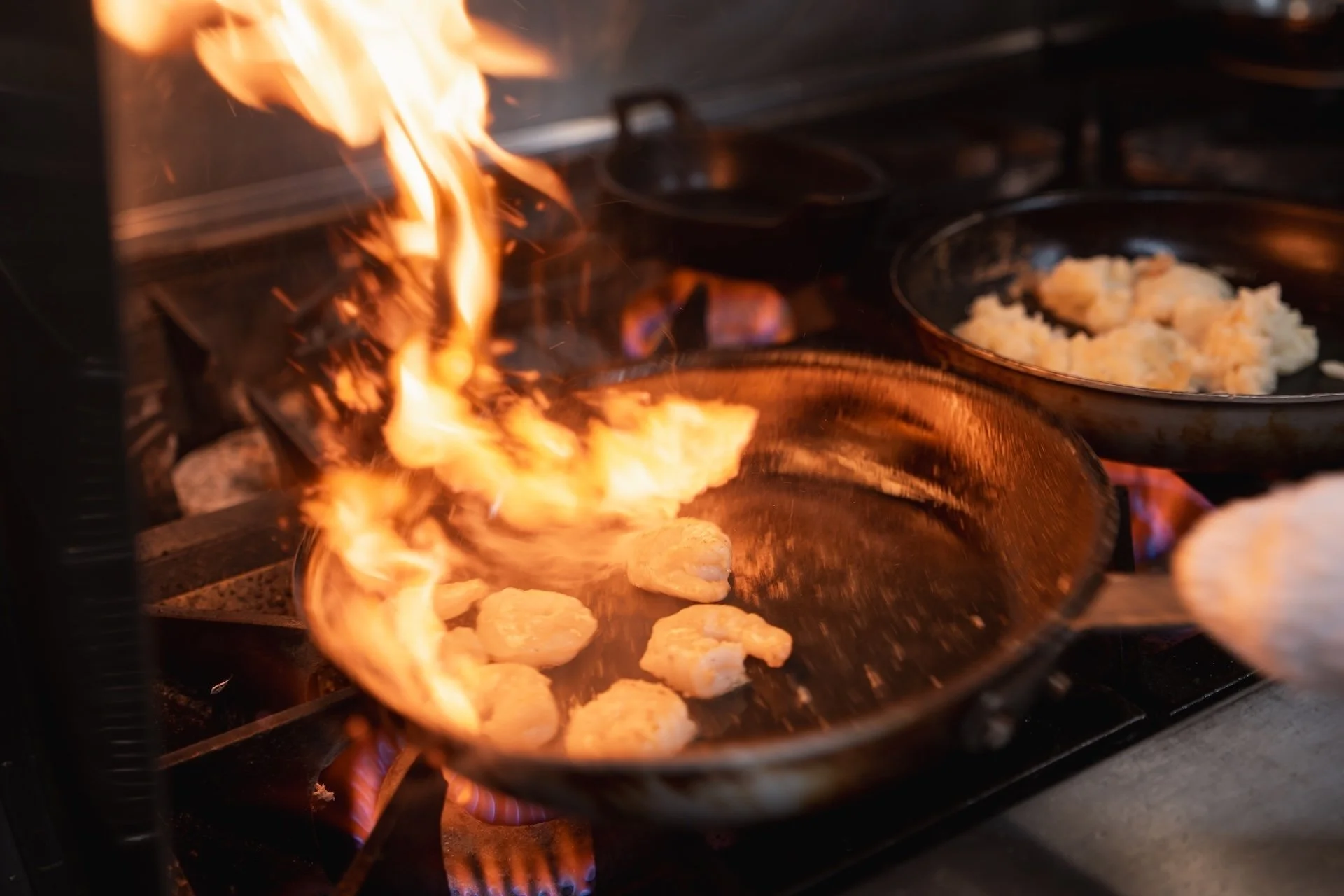 Chicken pieces cooking in a sizzling pan with flames rising, multiple pans with mashed potatoes in the background.