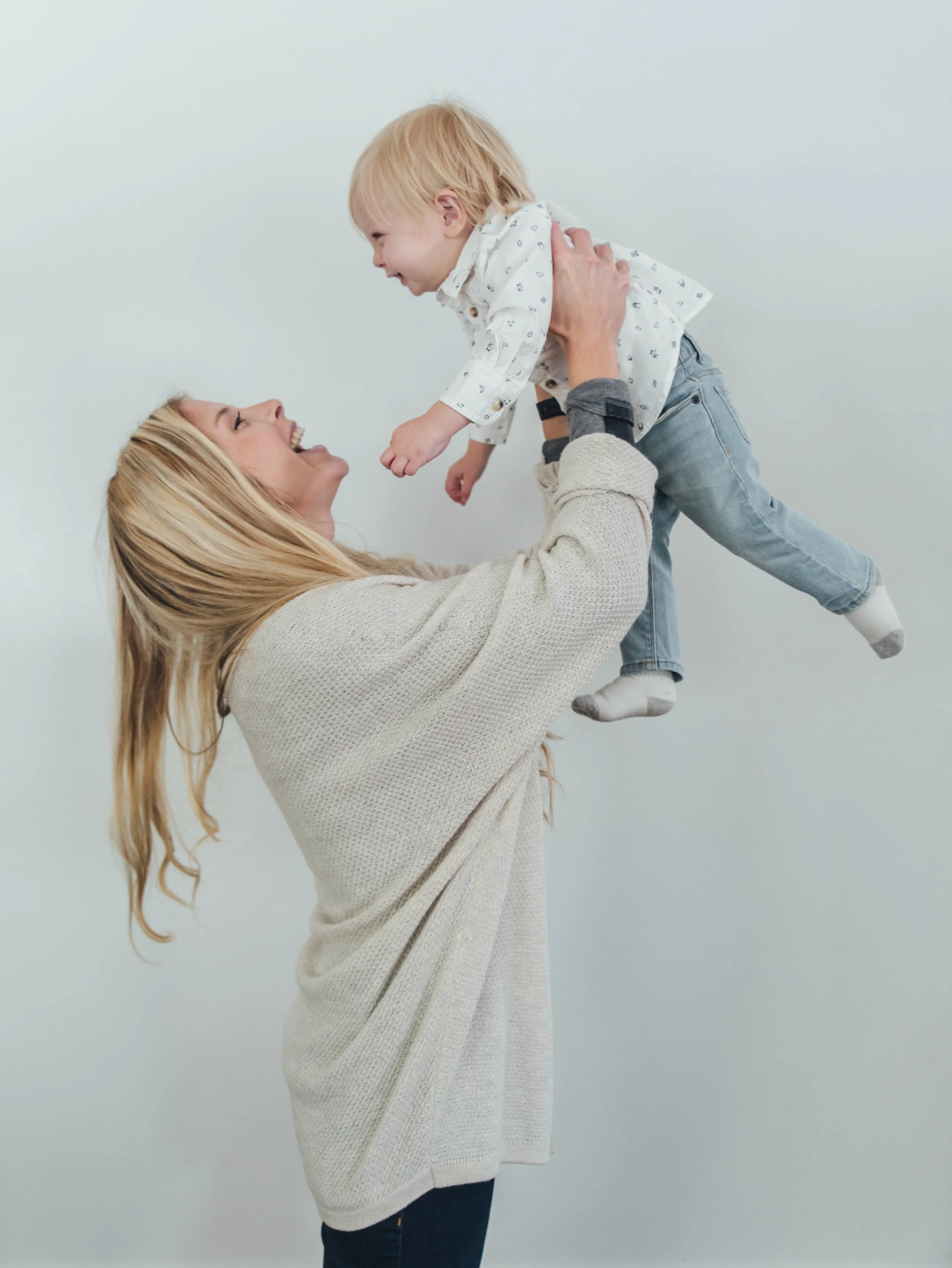 Woman lifting smiling toddler, both looking at each other, against plain background