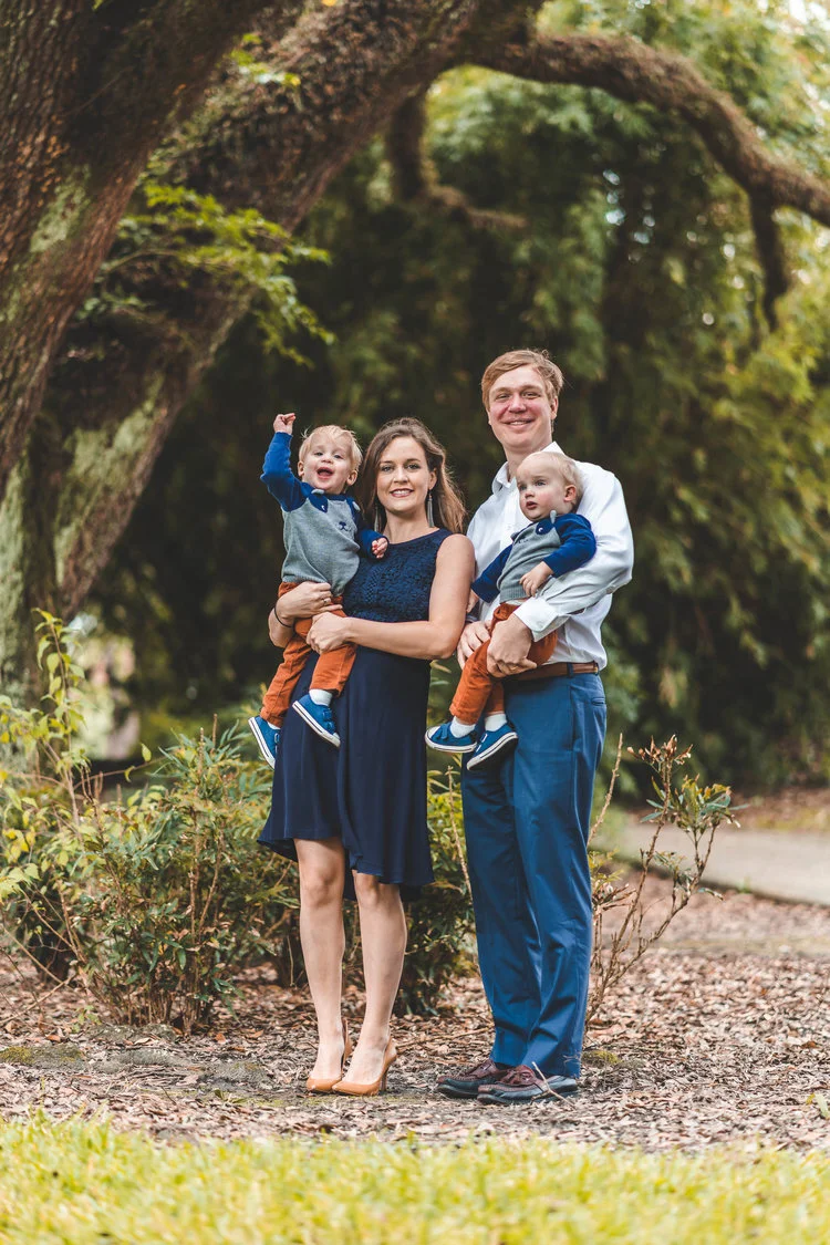 A family with two young children posing outdoors, under a large tree, while dressed in coordinating blue outfits.