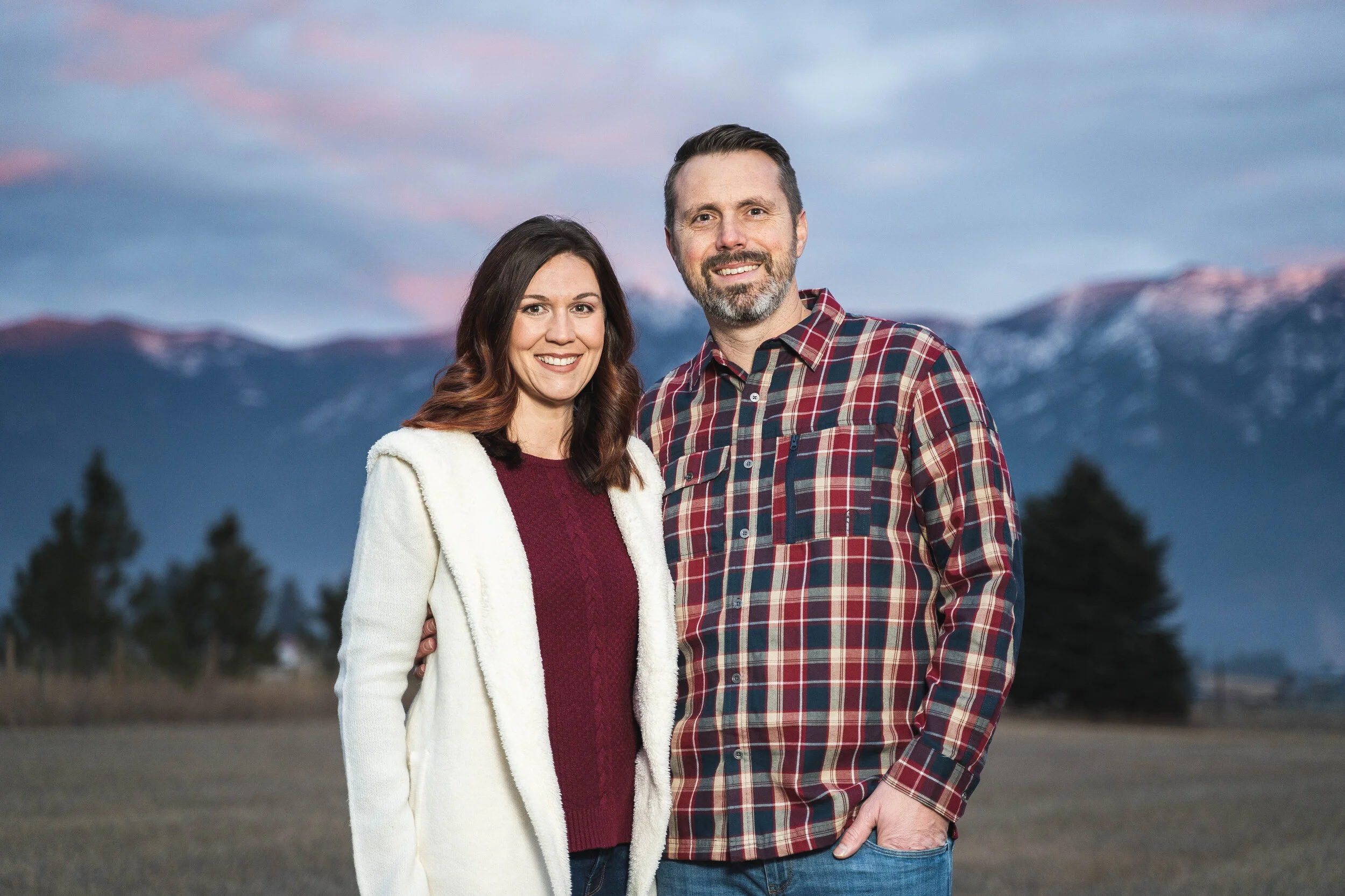 A man and woman standing outdoors with mountains in the background, wearing casual clothing.
