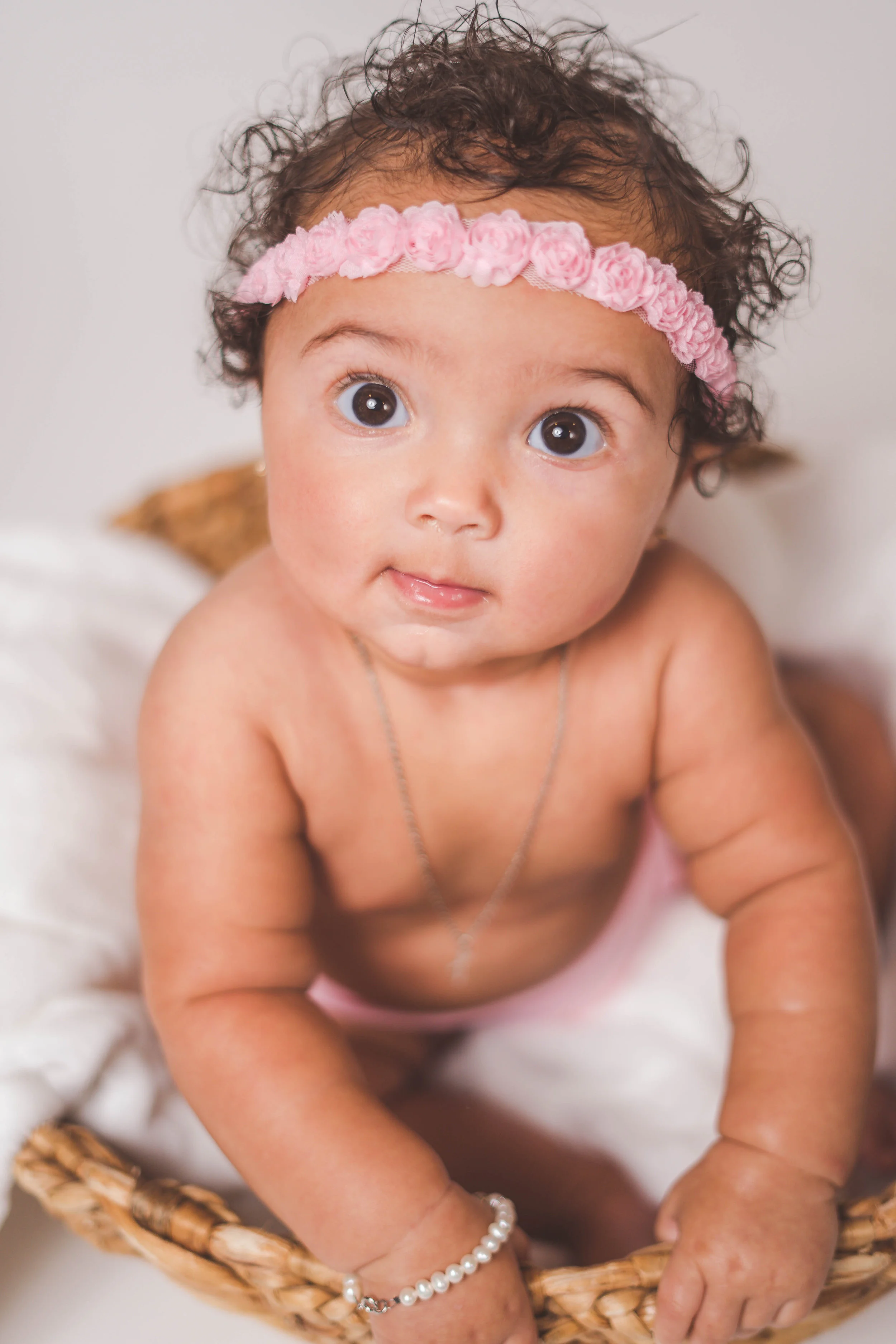 A baby with curly hair wearing a pink flower headband and pearl bracelet in a wicker basket.