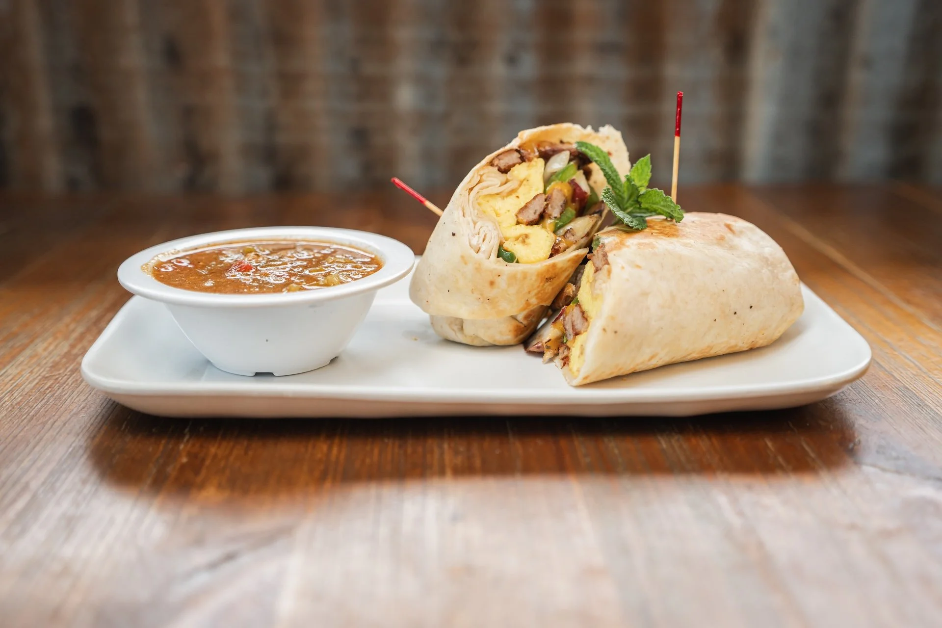 A plate with two cut wraps filled with meat, vegetables, and eggs, garnished with a sprig of mint, served with a small bowl of brown sauce on a wooden table.