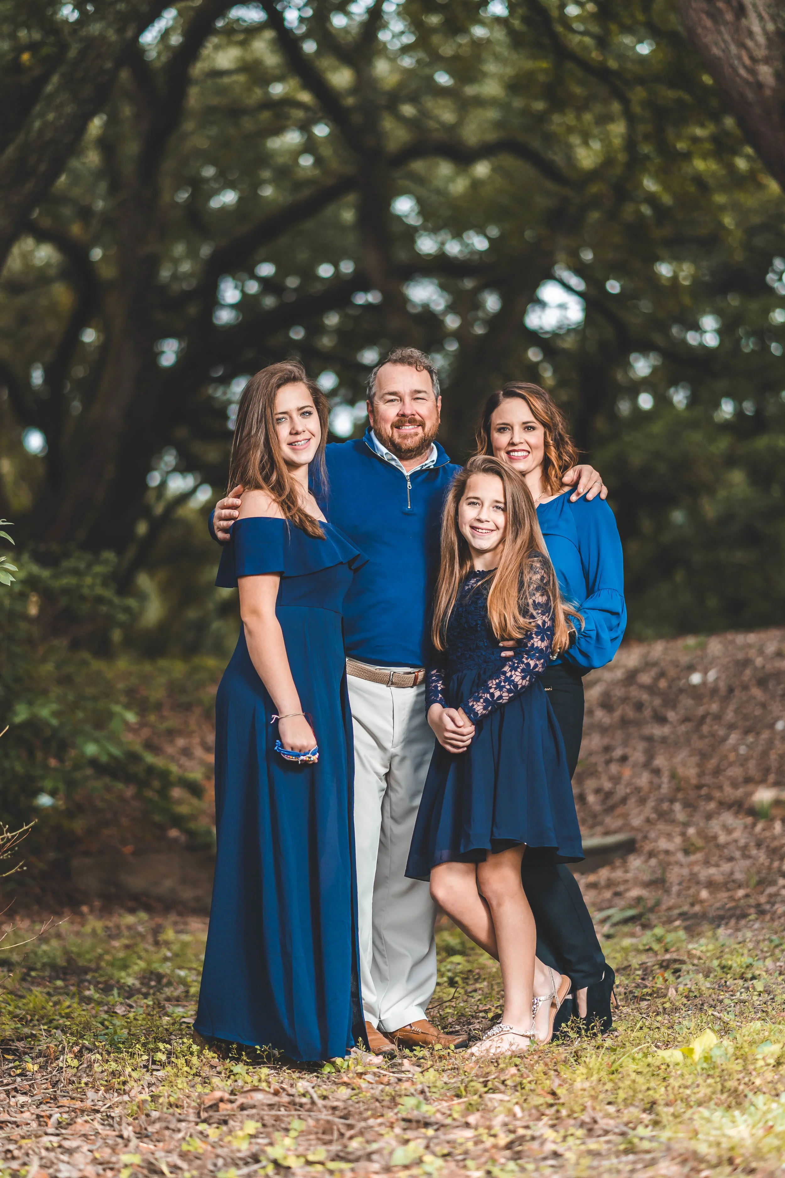Family of four smiling outdoors, dressed in blue, under trees.