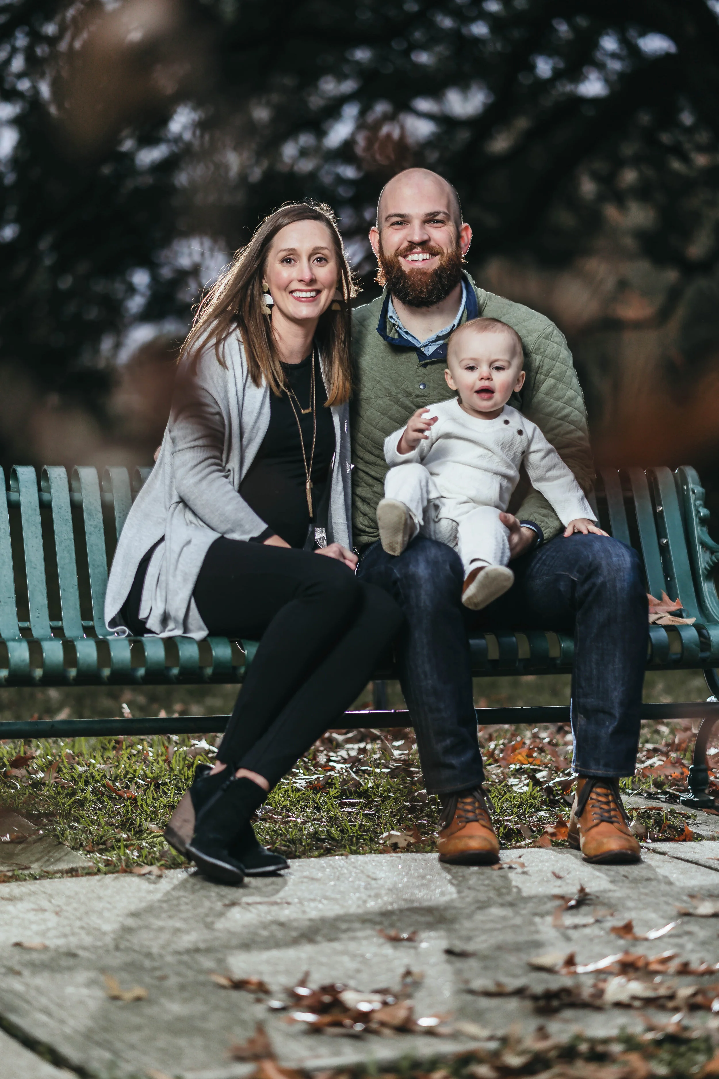 A family of three on a bench in a park, with parents sitting and smiling, holding a baby. The background features trees and fallen leaves.