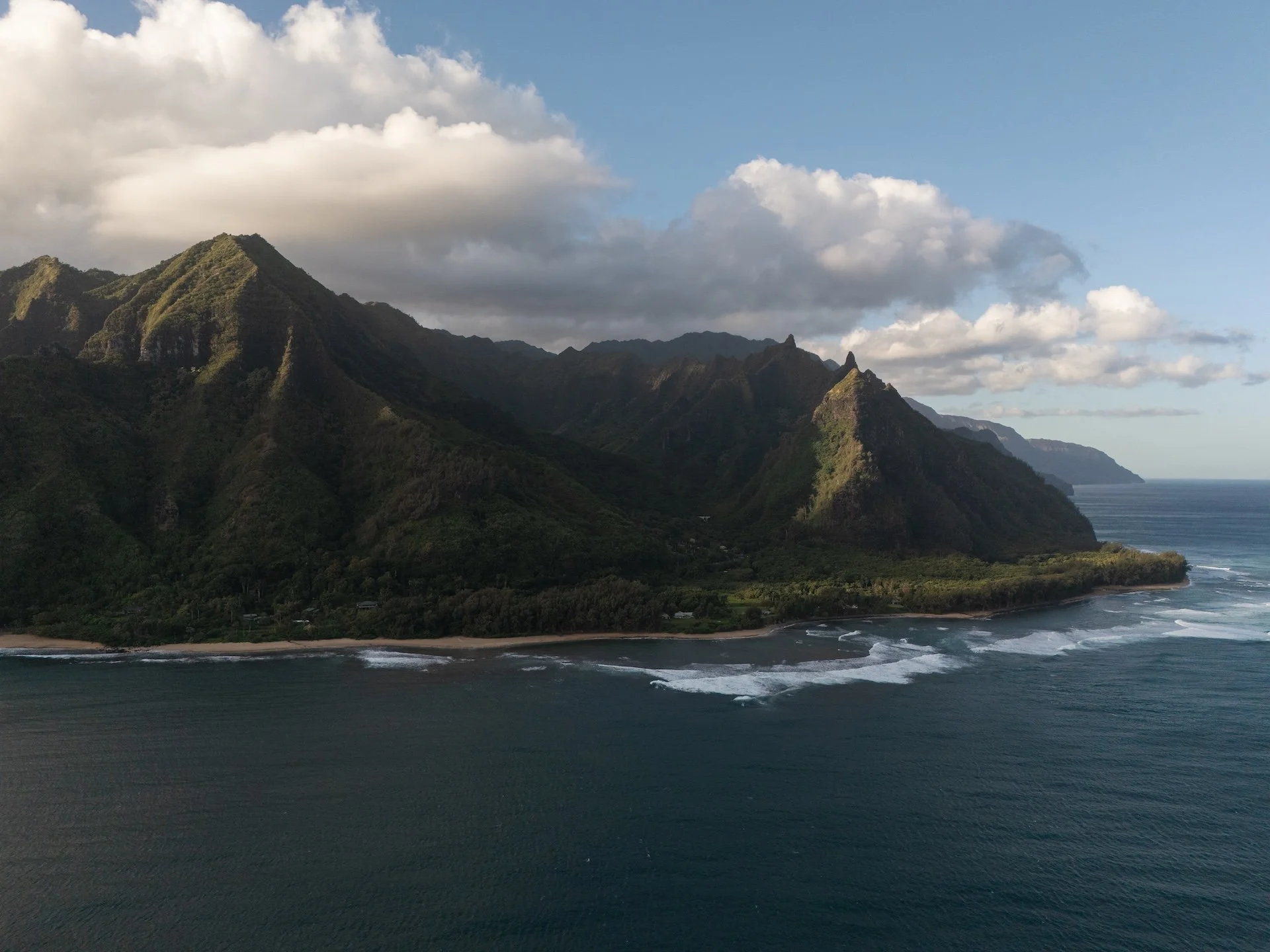 Aerial view of lush green mountainous island with rocky peaks, surrounded by ocean with waves and a partly cloudy sky.