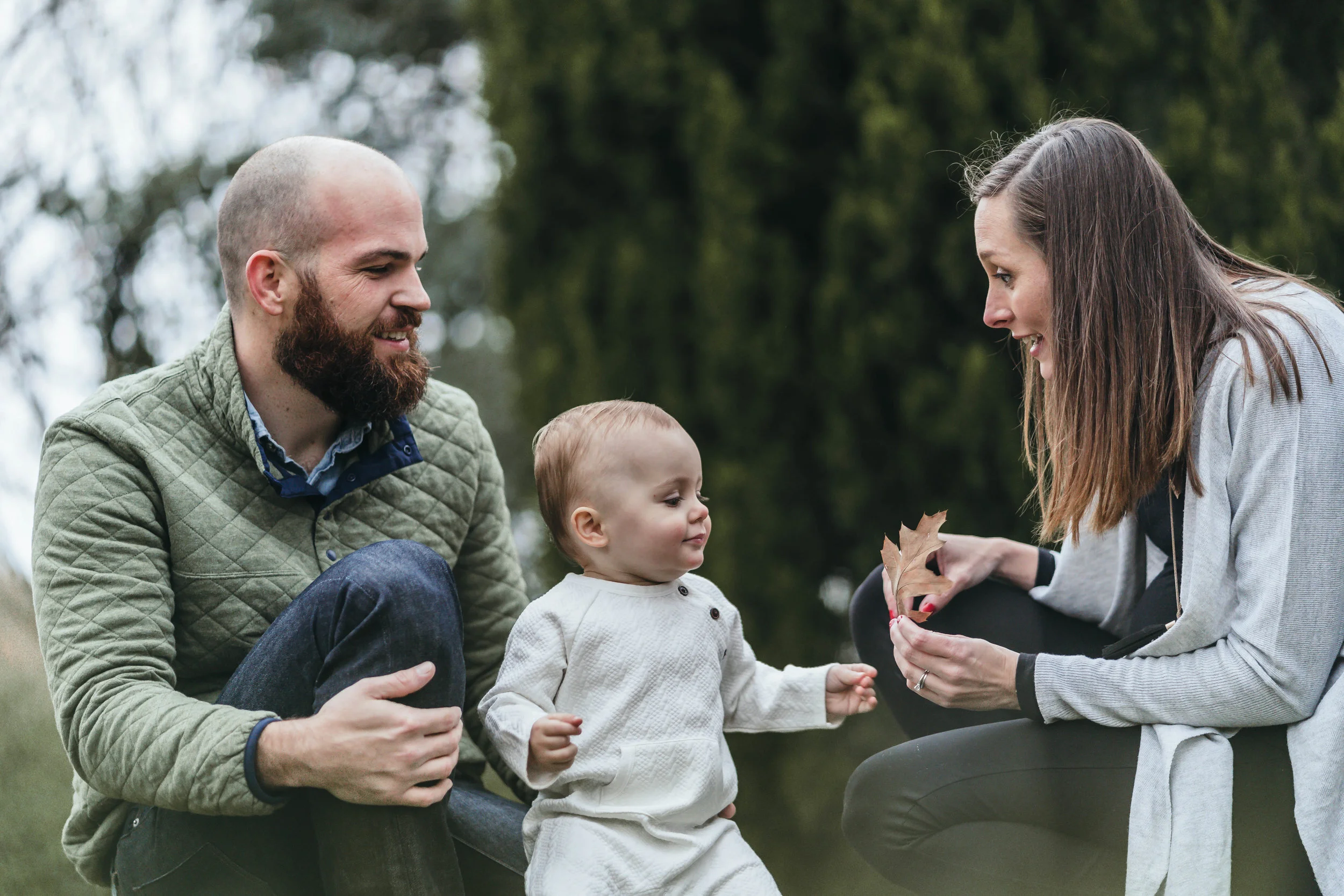 A family outdoors, with a bearded man in a green jacket, a baby in a white outfit, and a woman holding a leaf, smiling and interacting.
