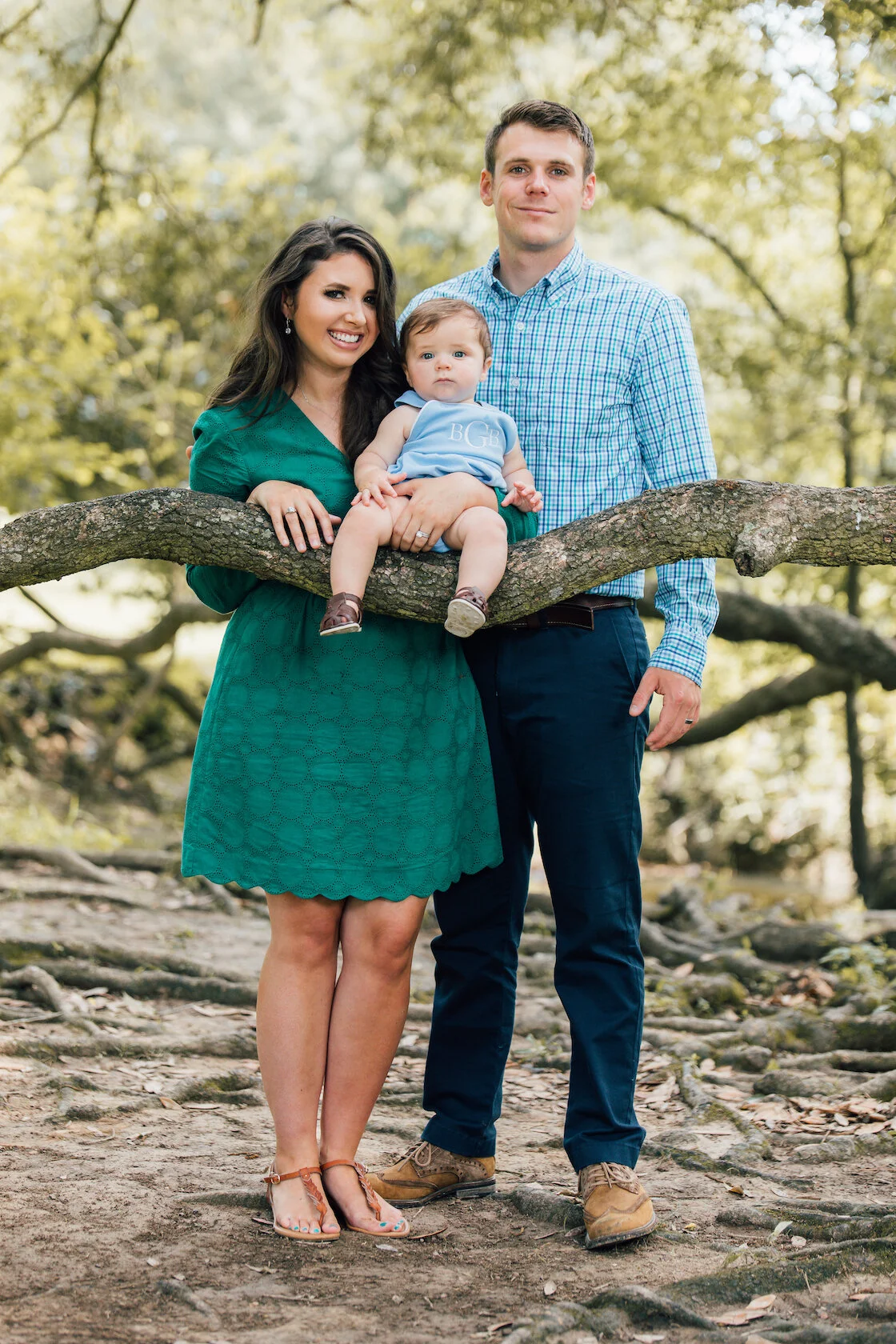 A smiling woman in a green dress and a man in a blue checkered shirt stand outdoors on a dirt path holding a baby dressed in light blue, sitting on a tree branch. The scene is a wooded area with trees and scattered roots.