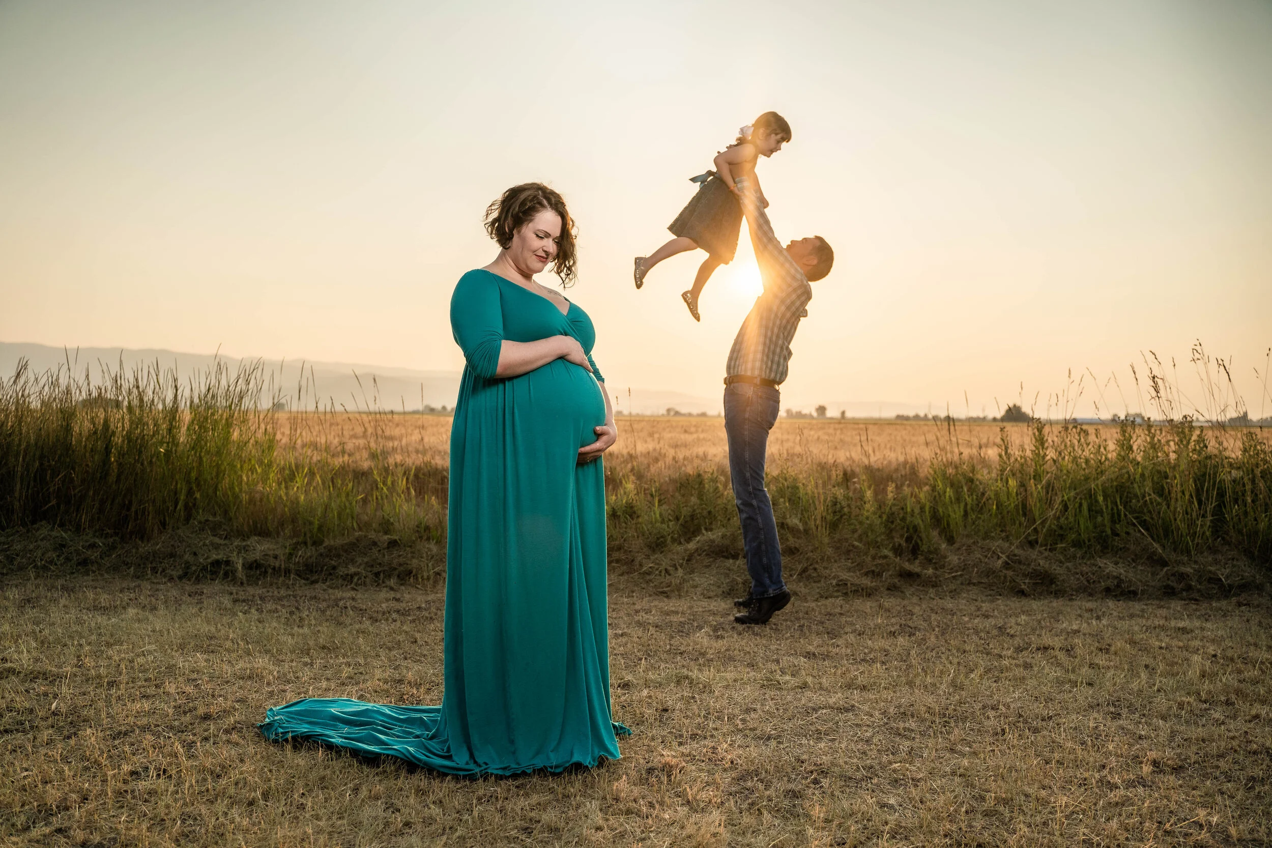 Pregnant woman in a teal dress standing in a field, smiling and cradling her belly. A man is lifting a child in the air, with the sun setting in the background.