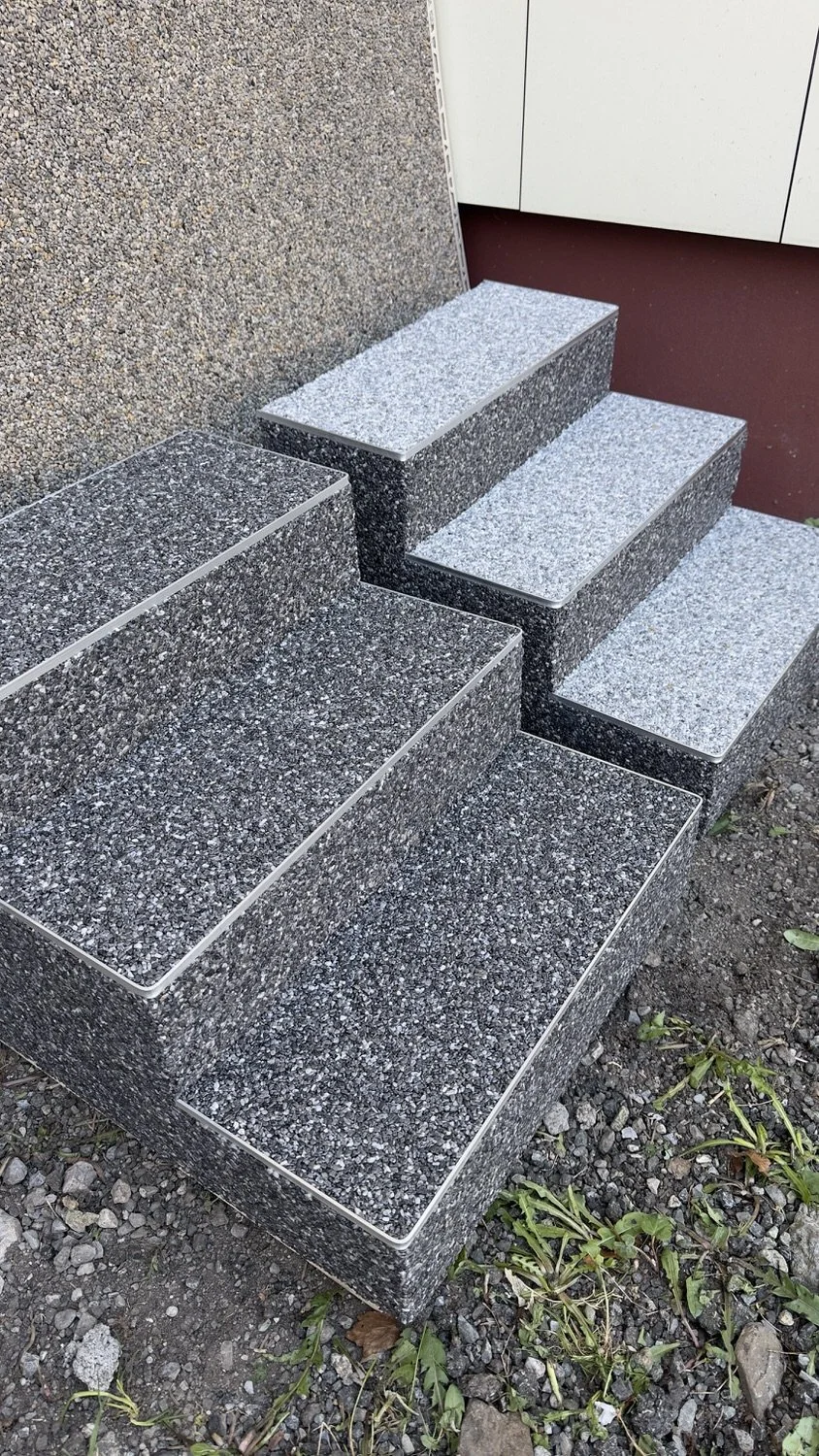 Gray speckled stone stairs next to a textured wall and gravel ground with scattered green plants.