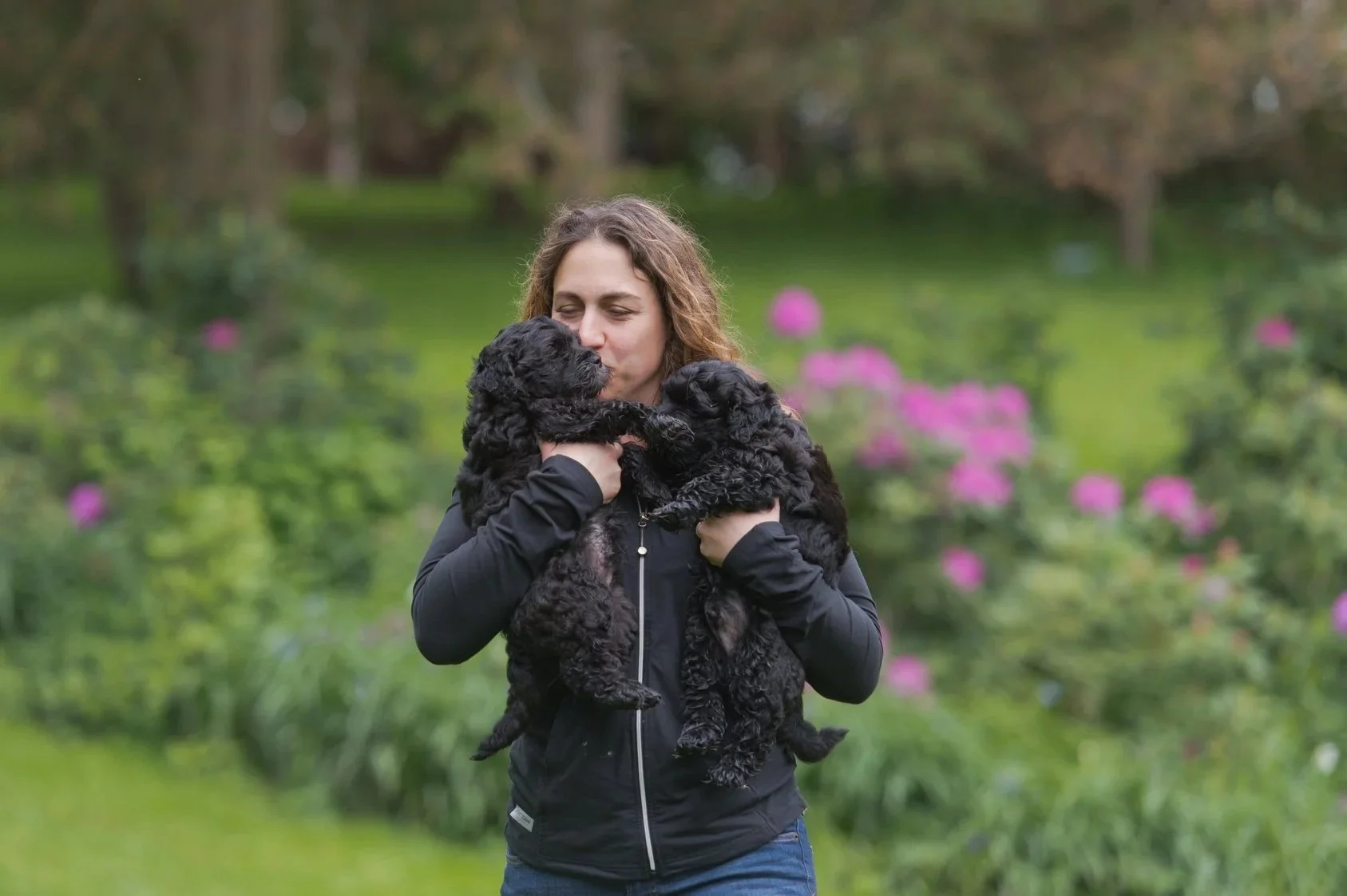 A woman holding two black puppies outdoors in a garden with pink flowers and green trees.