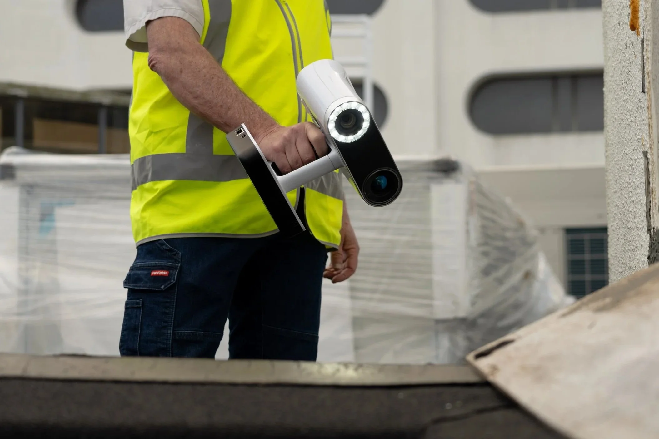 Person wearing a yellow safety vest holding a handheld 3D scanner at a construction site.