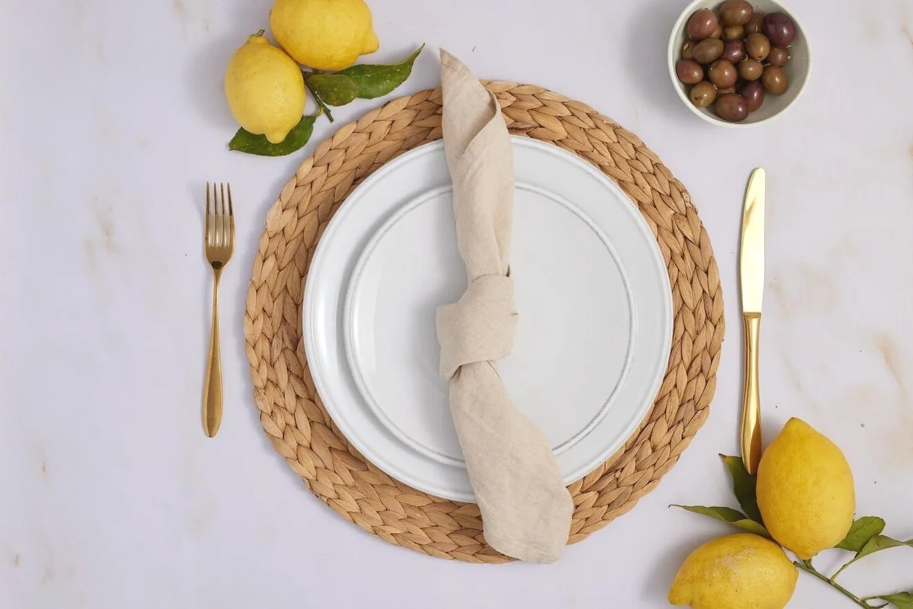 Empty white plate with a beige cloth napkin tied in a knot, placed on a woven placemat, with gold fork and knife on a white tablecloth. Lemon and leaf decoration at the top left corner, and a bowl of olives at the top right corner.