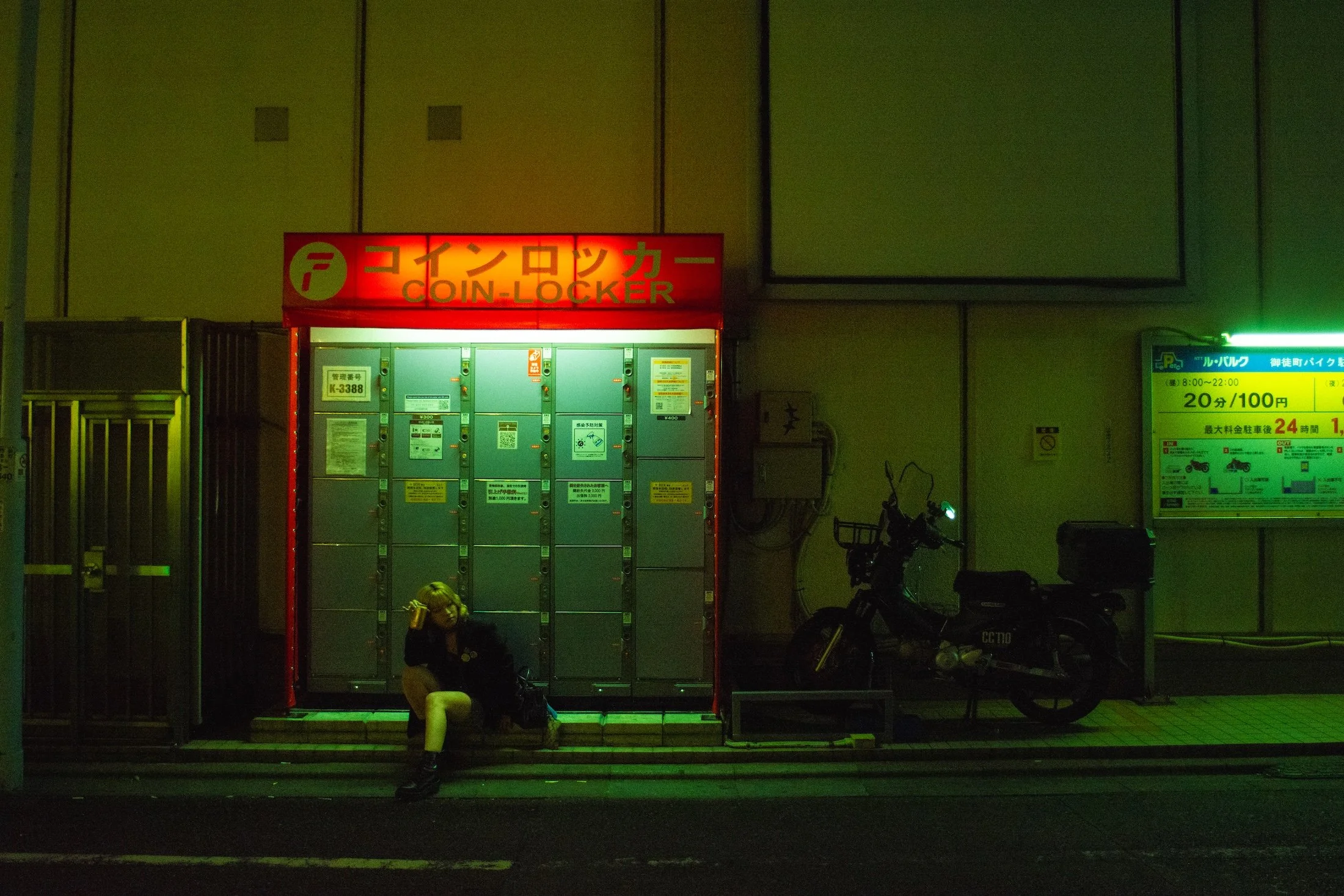 Nighttime scene of a coin locker station with a woman sitting on the steps, looking at her phone, and bicycles parked nearby. There are illuminated signs with Japanese and English text, including the words 'COIN-LOCKER' in red.