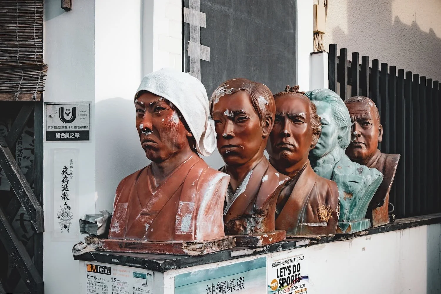 Bust sculptures of famous Asian leaders with some paint or paper on their faces, lined up on a platform outside a building, with Japanese and English text on signs nearby.