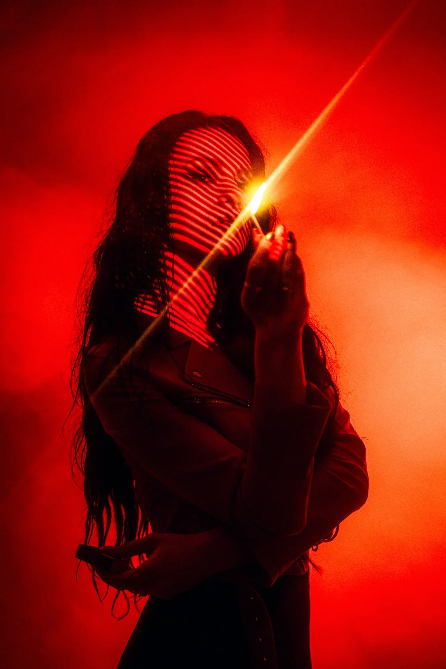A woman with long, curly hair holding a cigarette in her right hand, illuminated by a bright light, with red and orange hues creating a silhouette effect.