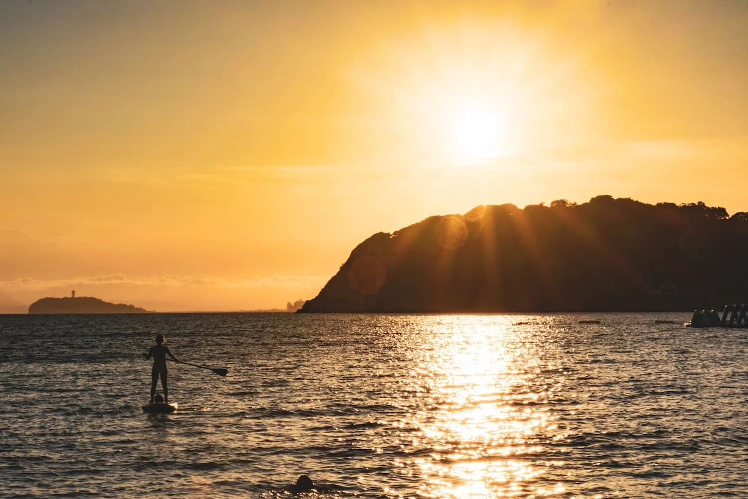 A person paddleboarding on the ocean during a sunset with a hill in the background.