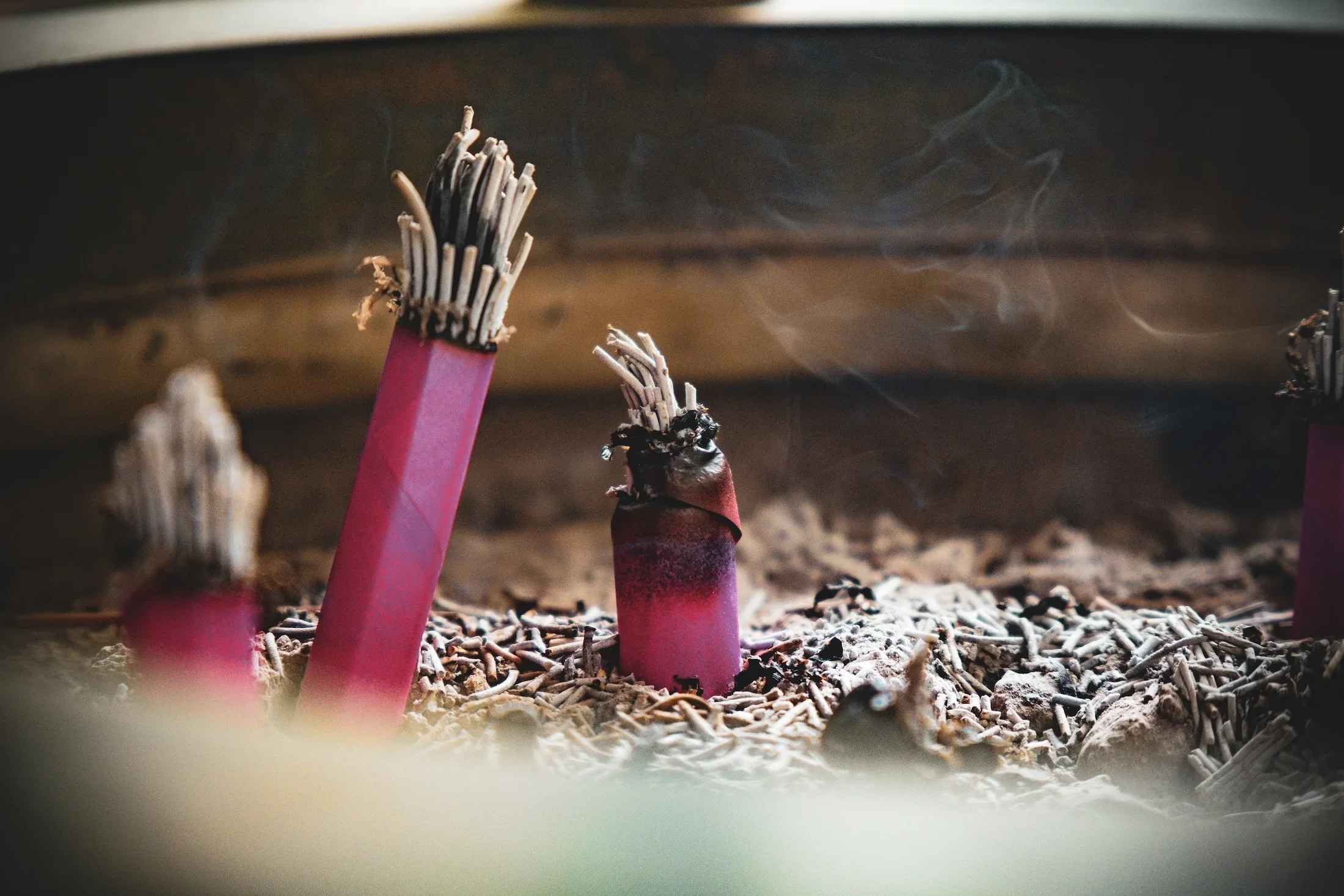 Several pink incense sticks are burning and releasing smoke, with ashes and small pieces laying around them, inside an incense holder.