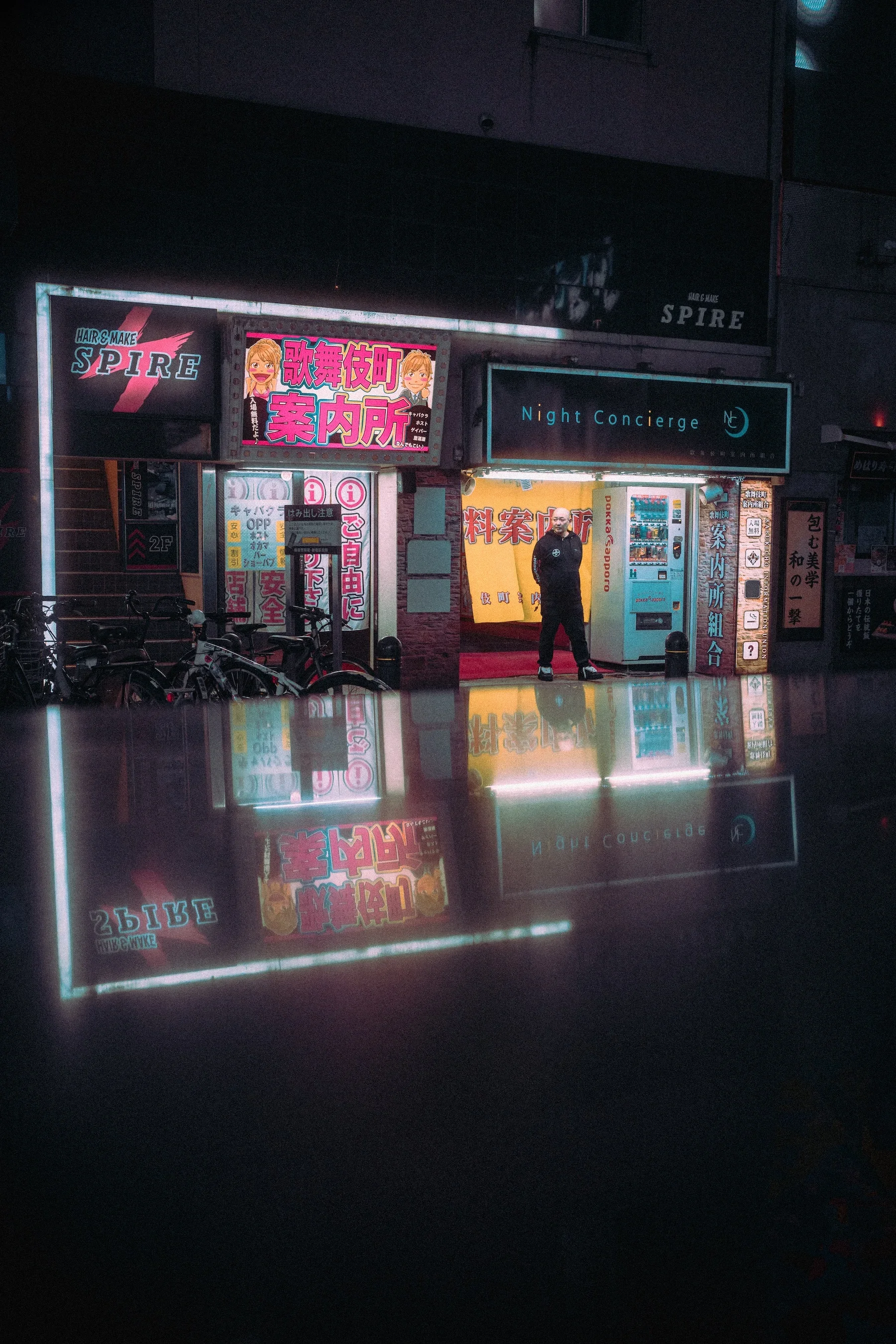 Night scene of a building with signs, including 'Hair & Make Spire' and 'Night Concierge,' with a security guard standing near the entrance, and bicycles parked outside, reflected in a glass surface.