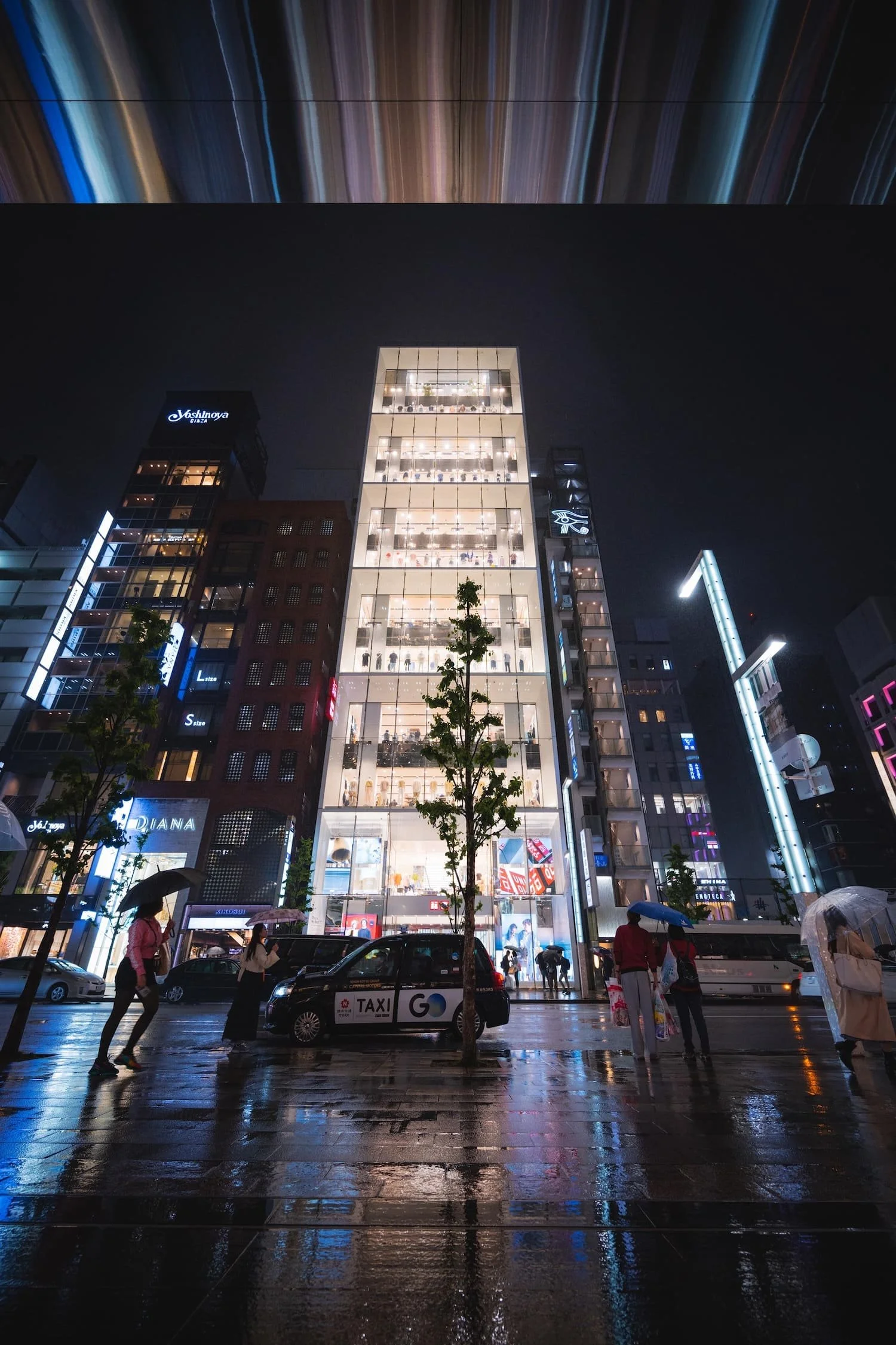 Night view of a city street with a tall, brightly lit glass building and pedestrians with umbrellas on a rainy evening.