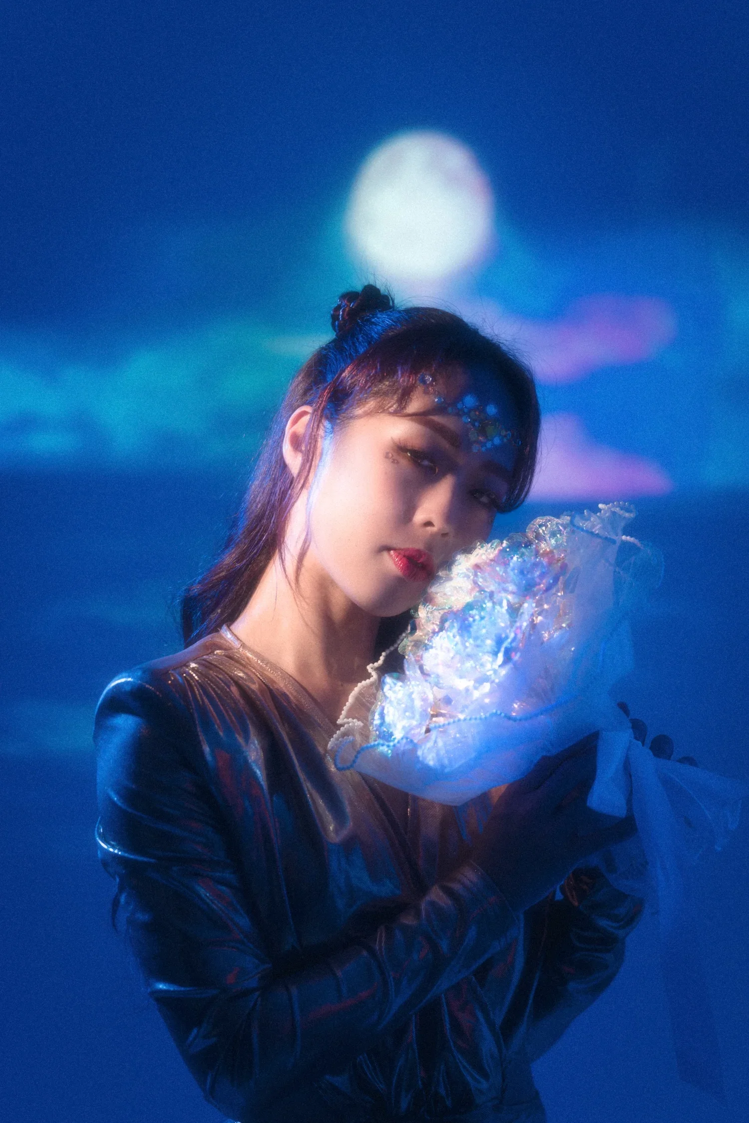 A young woman with dark hair in a bun and colorful makeup, wearing a shiny black jacket, holding illuminated bubble-like objects while standing in a dim, blue-lit environment with a large blurred light in the background.