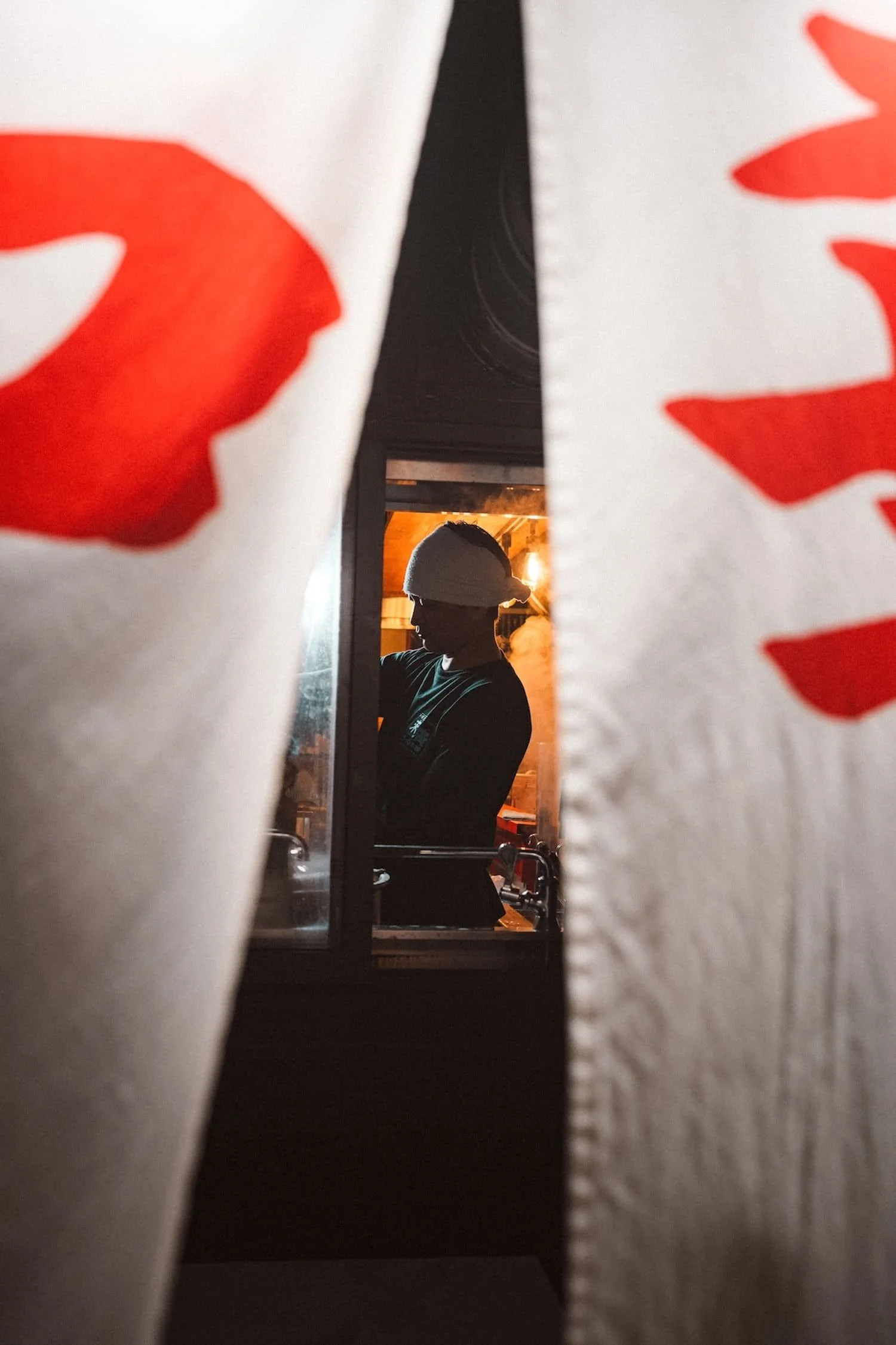 A person wearing a hat and black shirt seen through a narrow opening between two large white towels with red designs, with a warm indoor setting happening behind them.