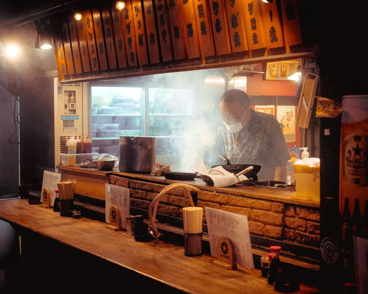 A chef cooking in a small Japanese ramen restaurant kitchen with smoke billowing, seen through a pass-through window. The restaurant has a wooden counter with menus, condiments, and utensils. Bright overhead lighting illuminates the scene.