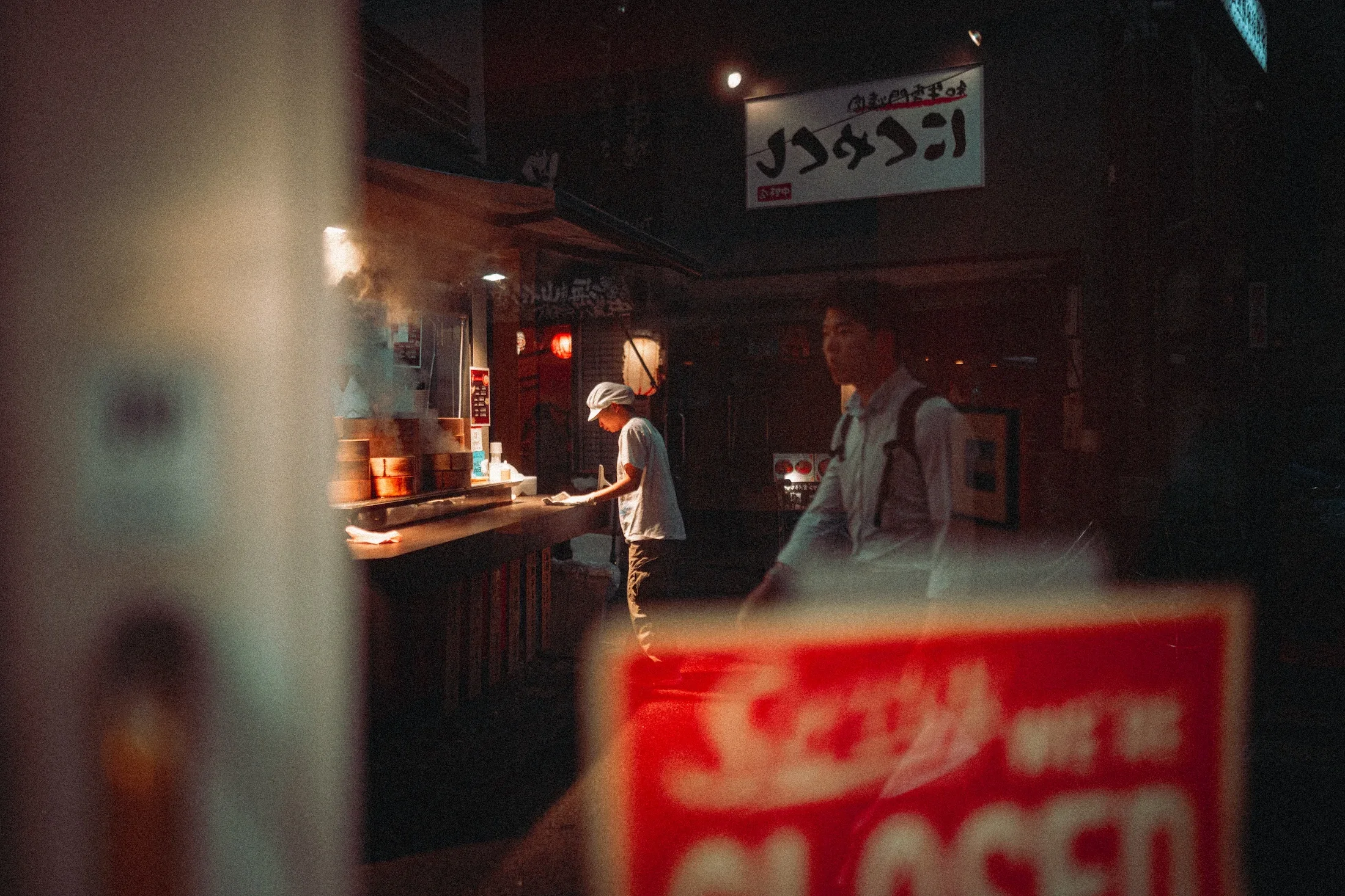 Blurred image of a restaurant or food stall with a chef working behind the counter and a person standing in front, with text in Japanese signs.