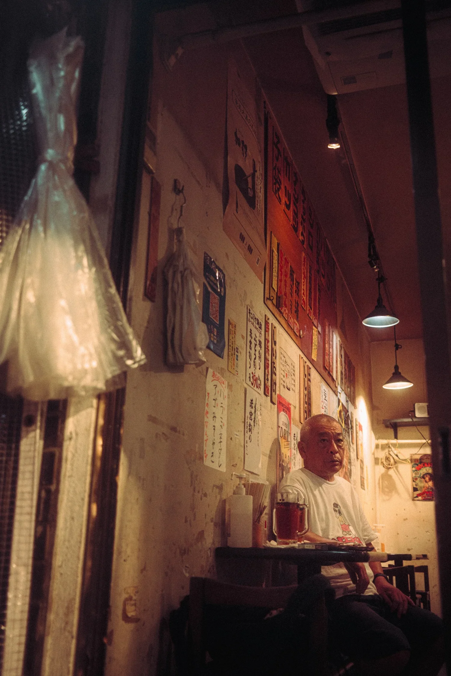 An elderly man is sitting inside a Japanese restaurant. The background features a wall decorated with various posters, menus, and signs in Japanese. The lighting is dim and warm, with hanging ceiling lights. The man appears to be waiting or resting, with a glass of beer in front of him.