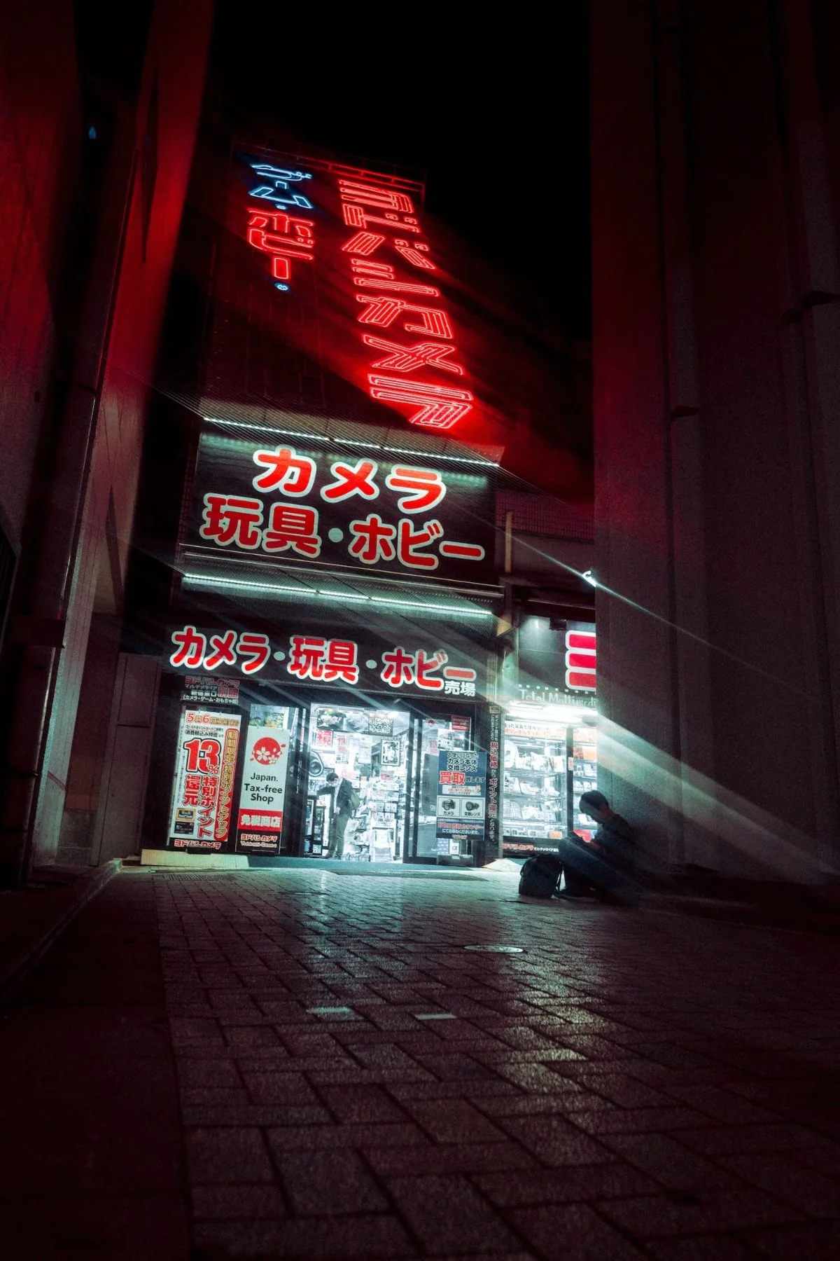 Nighttime view of a Japanese store with neon signs advertising cameras, toys, and hobbies, with a person sitting on the sidewalk in front.