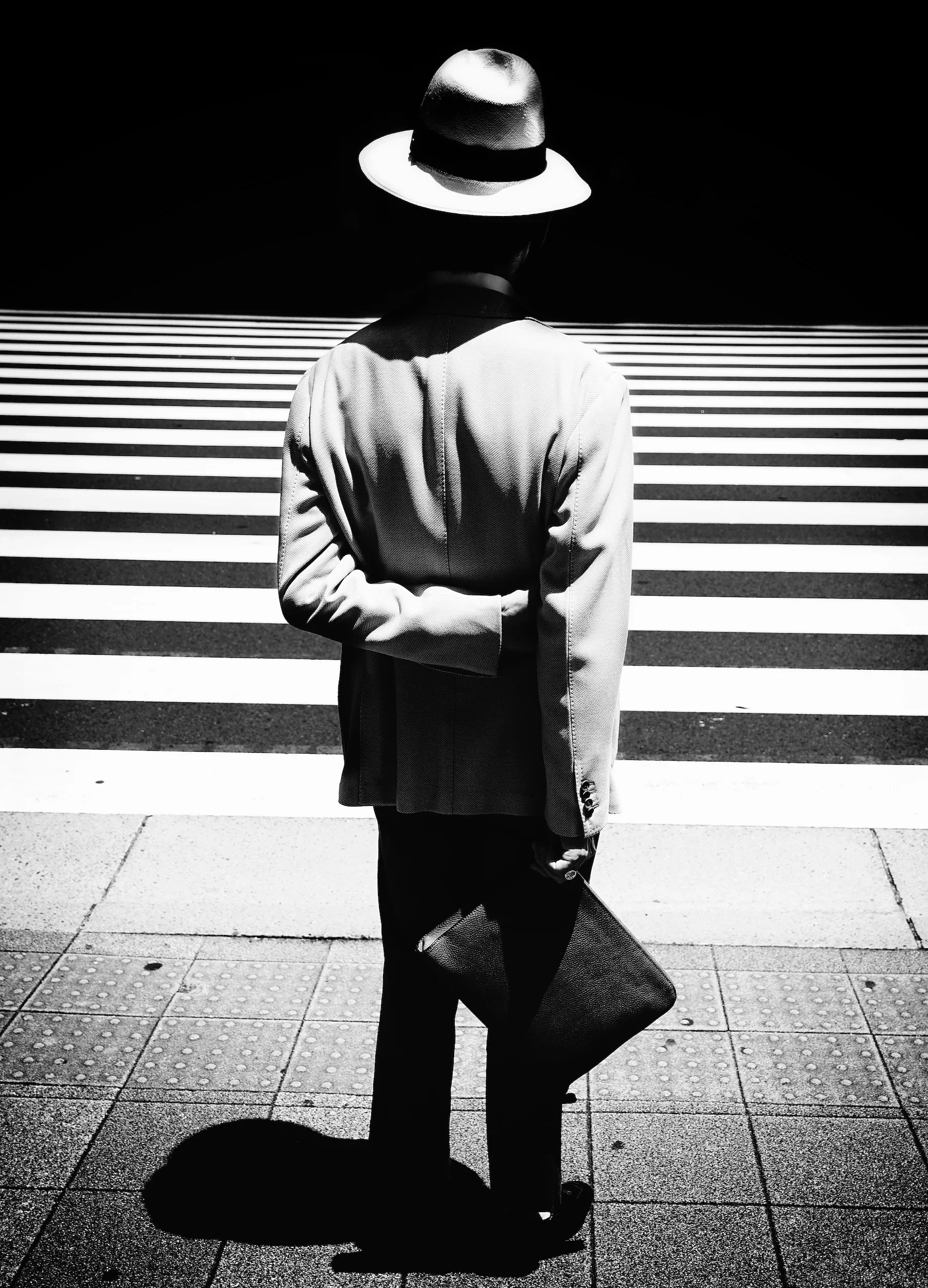 A person dressed in a blazer and hat, holding a bag, stands on a sidewalk under a crosswalk with striped lines.