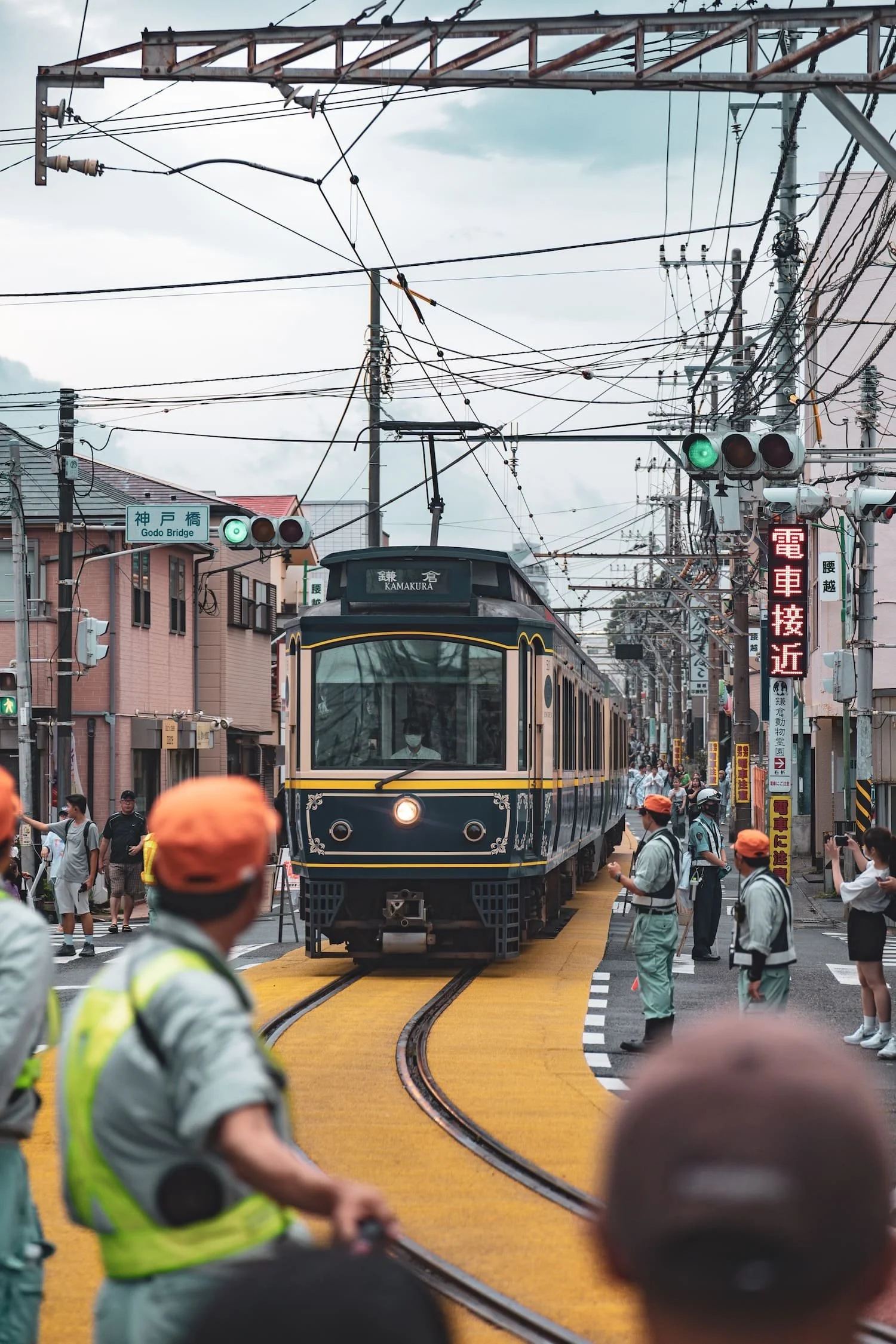 A vintage-style train with a black and cream-colored exterior traveling through a busy urban street with pedestrians and workers, overhead wires, and traffic signals.