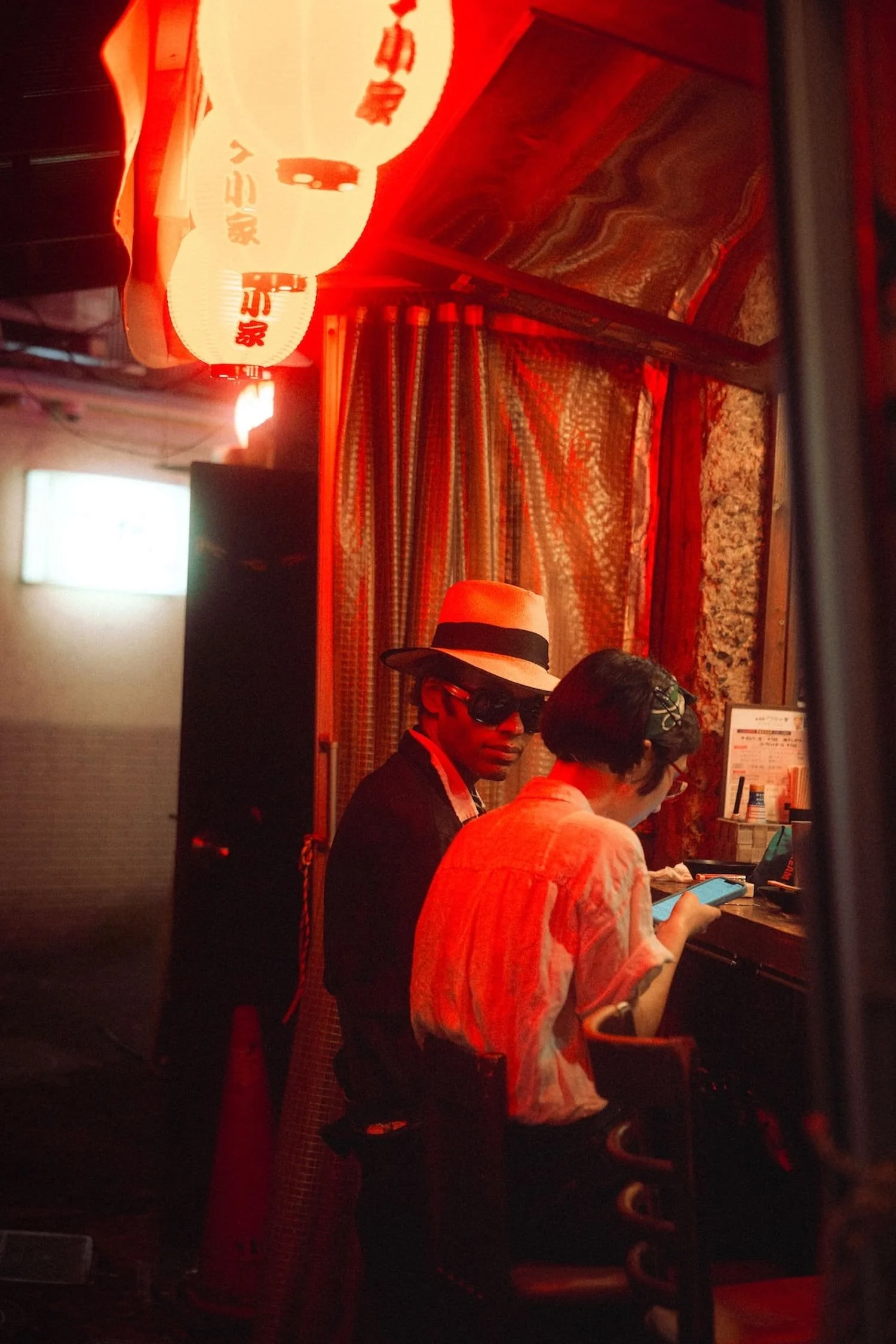 Two people sitting at a corner in a dimly lit space decorated with red neon lights and hanging lanterns with Asian characters. One person wears a white hat and sunglasses, while the other is looking at a phone.