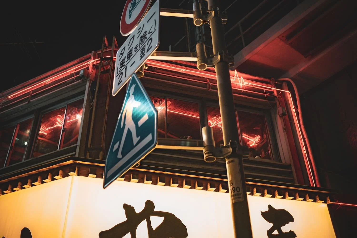 Nighttime city scene with street signs, a building with red neon lighting, and a lit billboard with black silhouette designs.